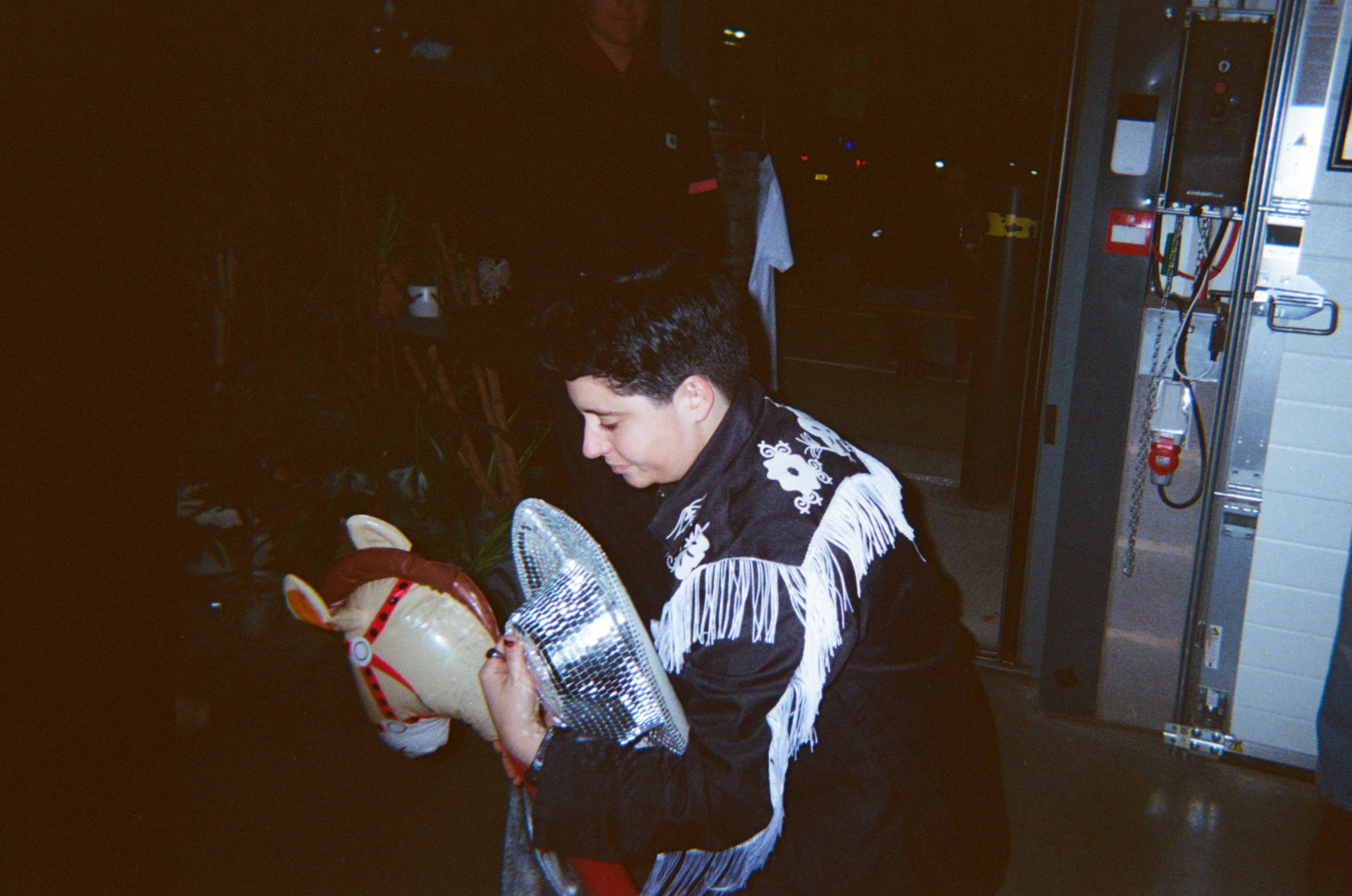 Woman in black patterned jumper holding baby in red and white outfit, sitting indoors with kitchen appliances visible in background.