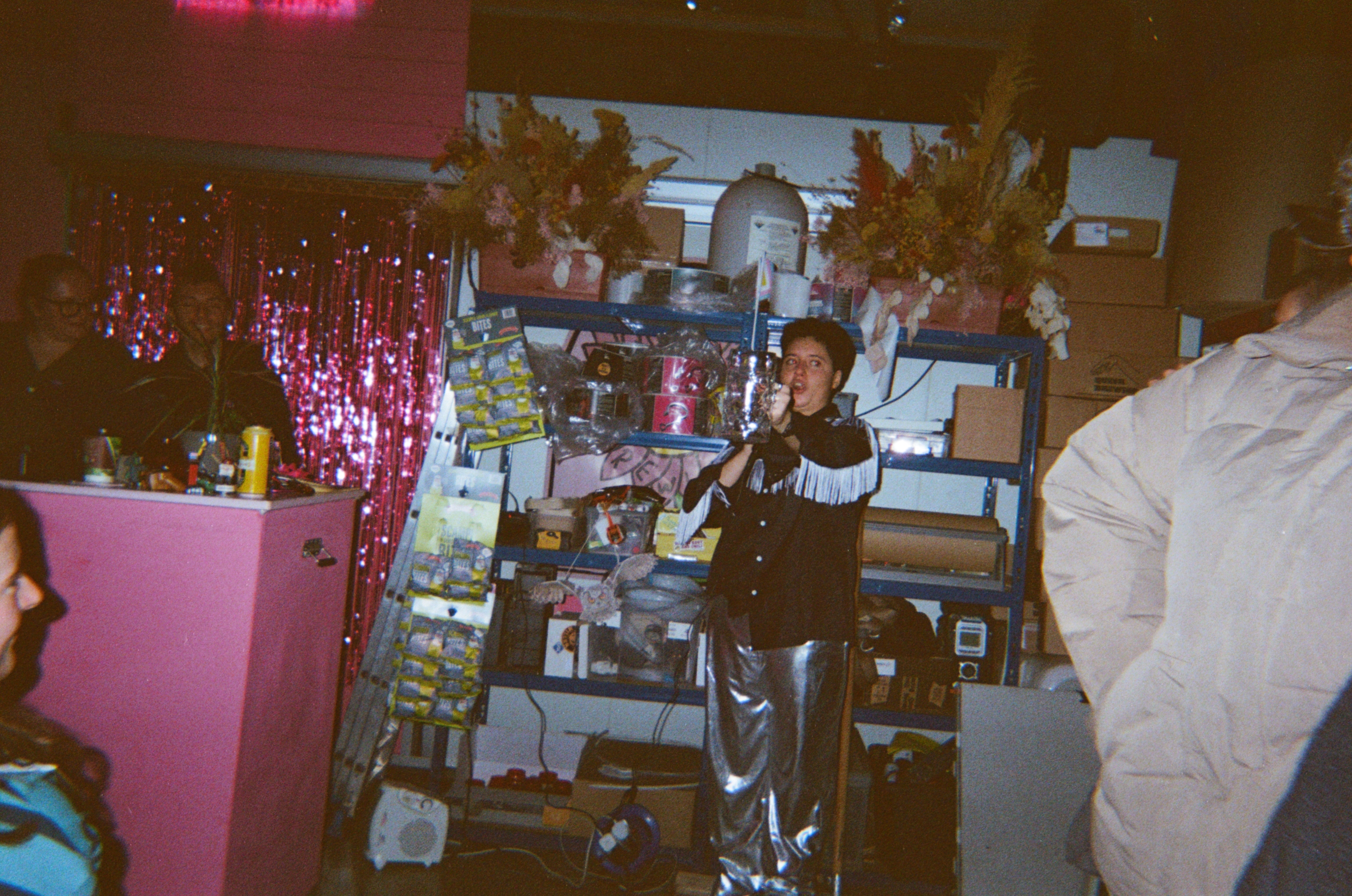 Person at microphone in cluttered room with pink surfaces, sequined curtain, shelving with items, and dim lighting.