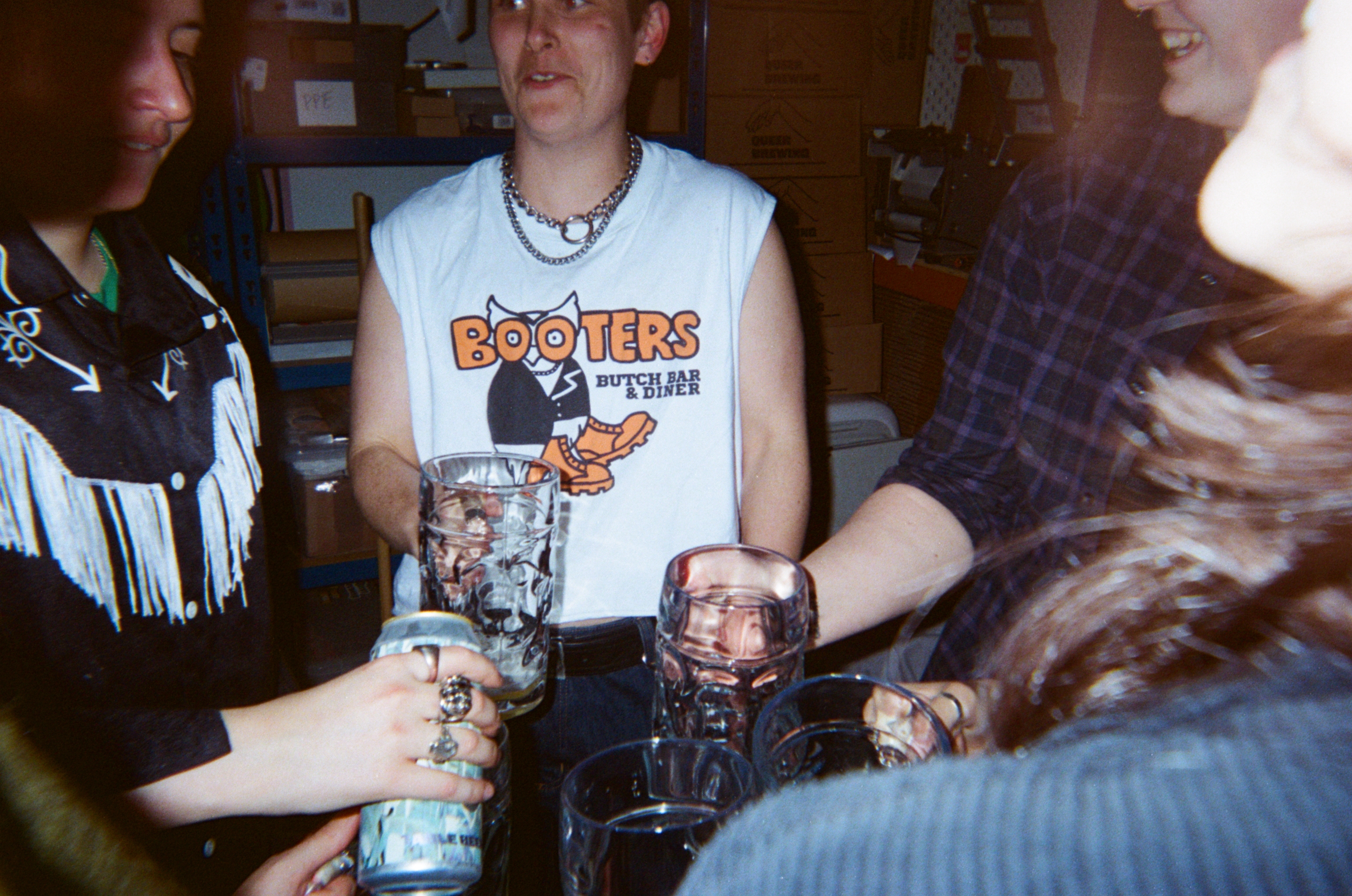 Group of young people sitting around table with drinks, one wearing Hooters vest, casual indoor setting with books in background.