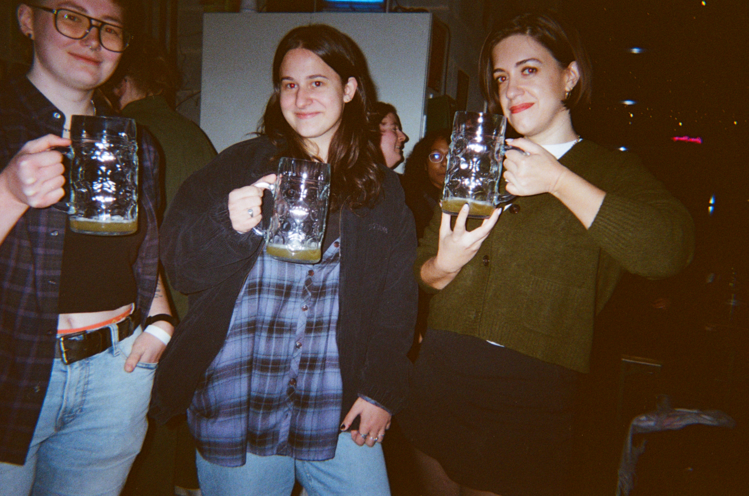 Three young women holding pint glasses in a dimly lit pub, wearing casual clothing including plaid shirt and dark tops.