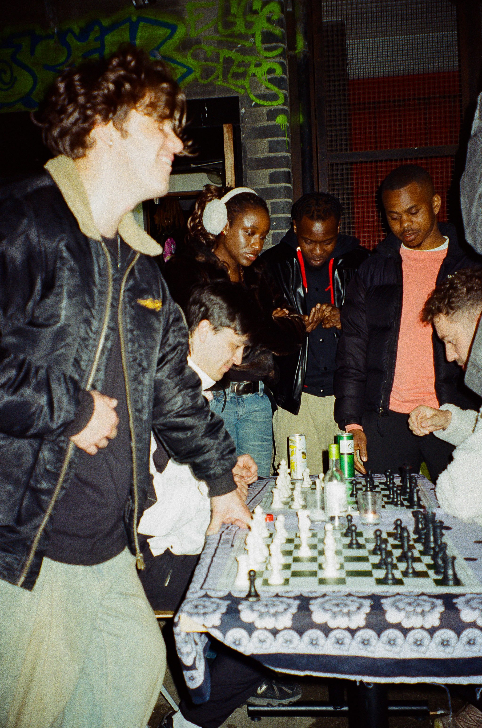 Group of people gathered around chess boards on street at night, green graffiti visible on brick wall background.