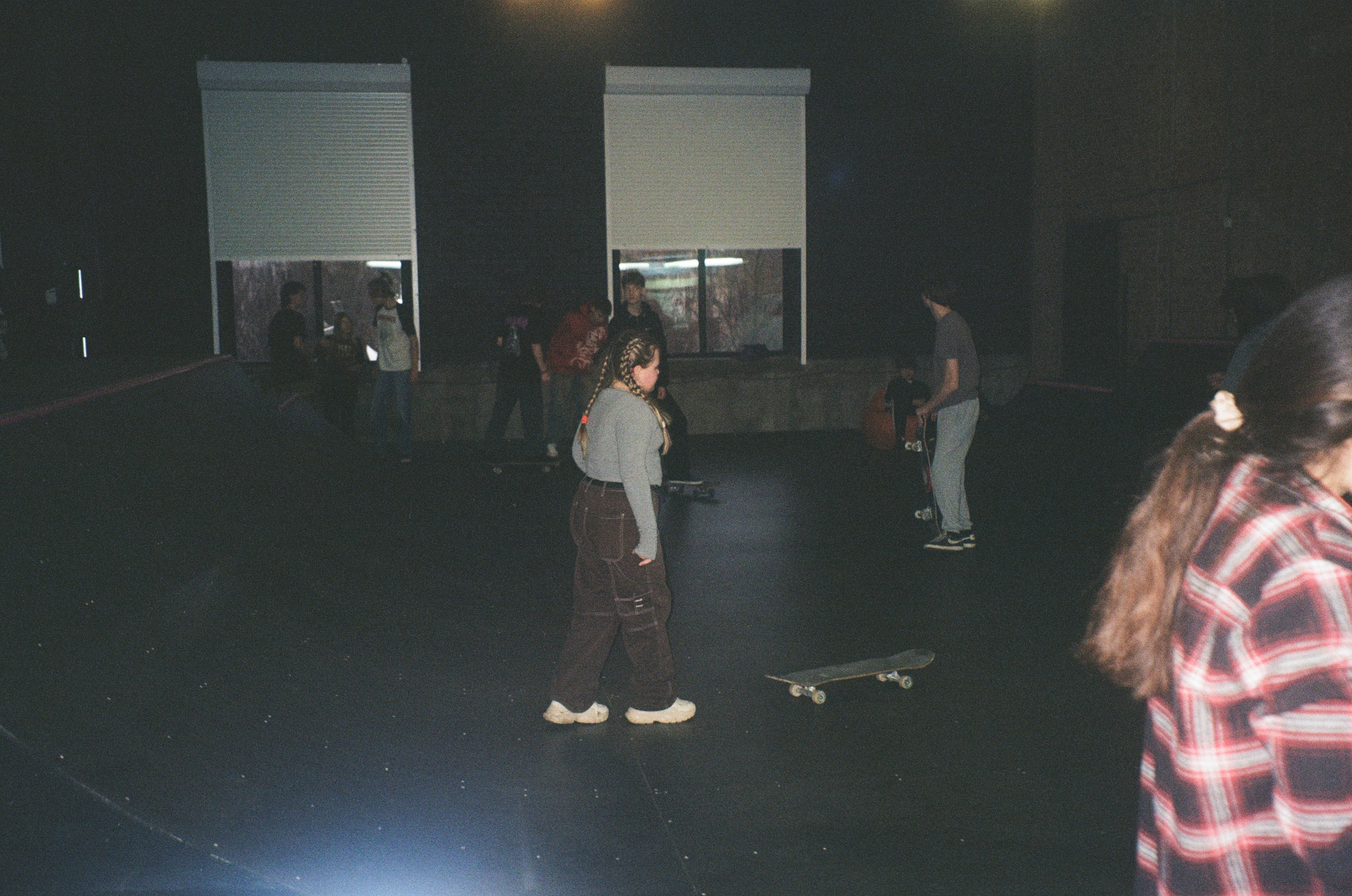 Dark indoor scene with people standing, some holding skateboards.