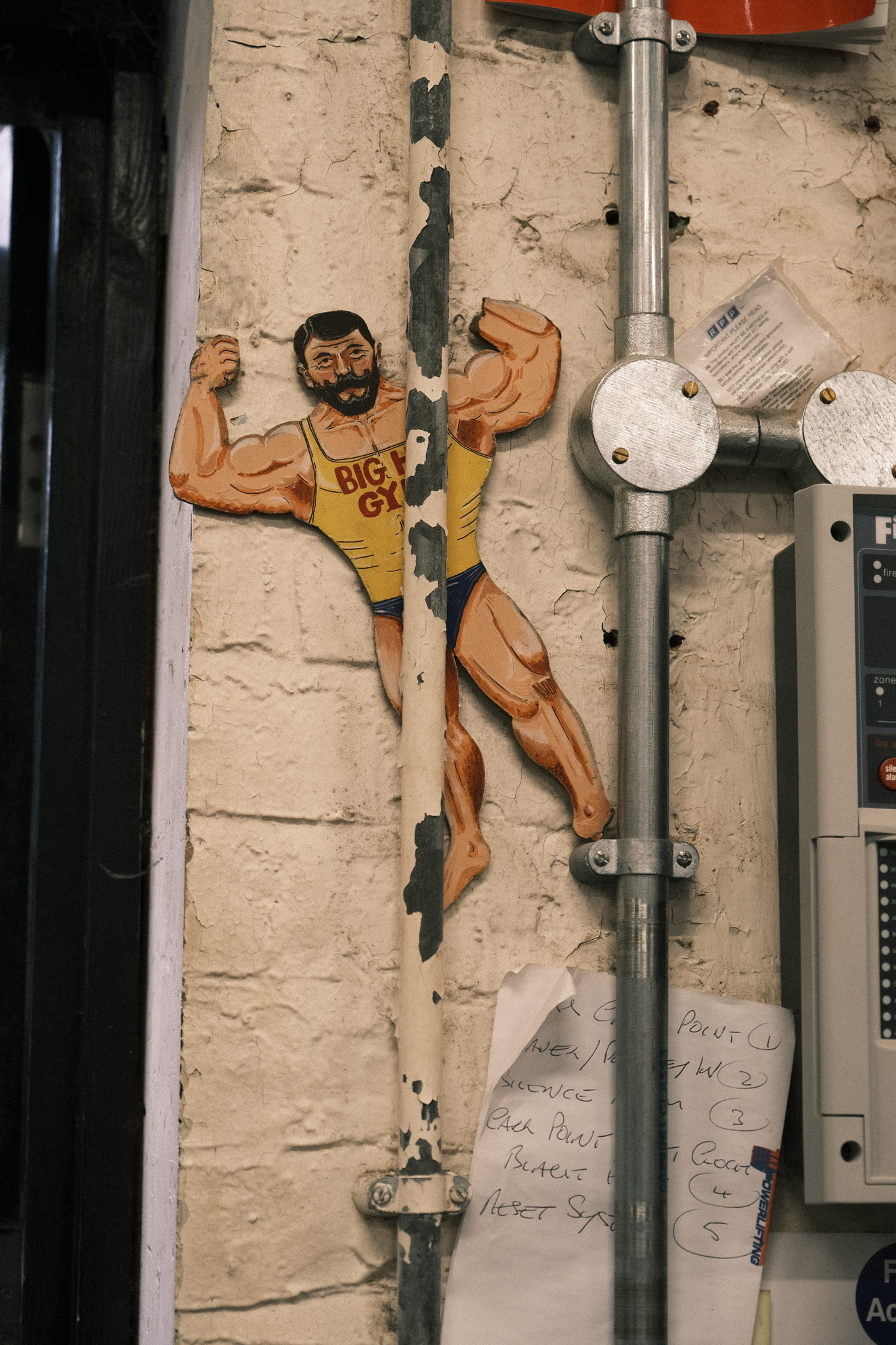 Cartoon strongman sticker on weathered cream wall next to grey electrical conduits and junction boxes with handwritten notes below.