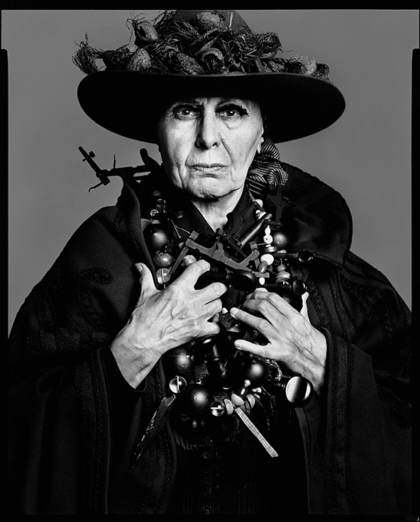 Elderly woman in ornate black hat and clothing holds decorative walking stick, black and white portrait with dramatic lighting.