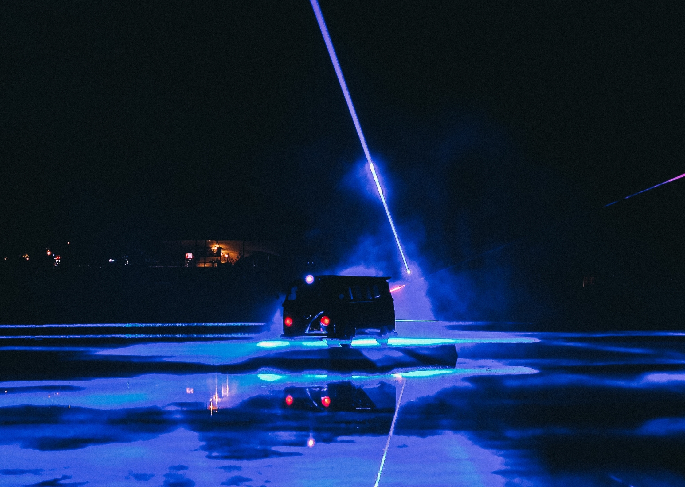 Dark vehicle on snowy ground with bright blue laser beam extending vertically upward, orange lights in background, wet surface reflections.