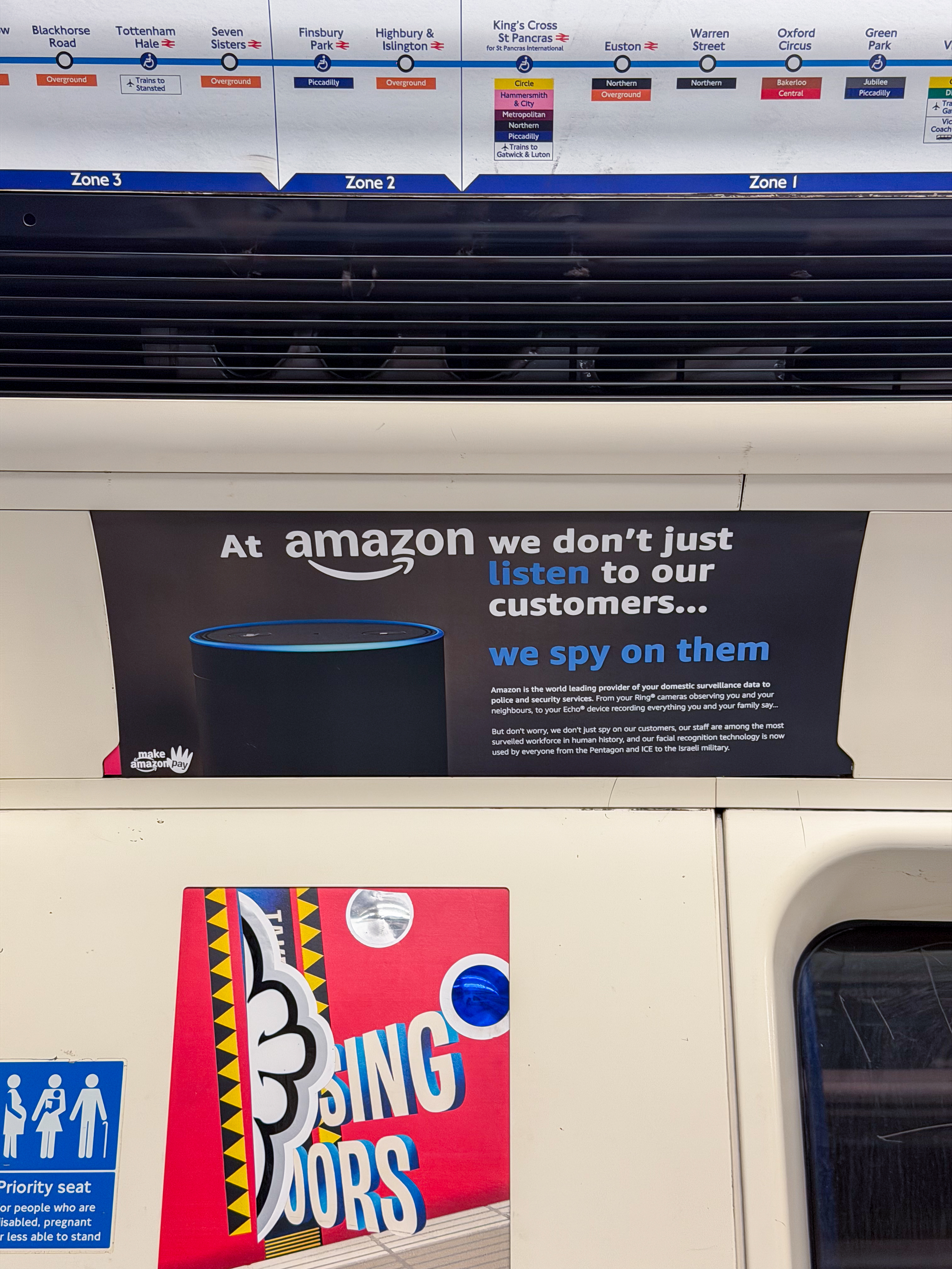 Underground station platform with zone map above, black Amazon advert with blue text, and red poster with yellow geometric patterns below.