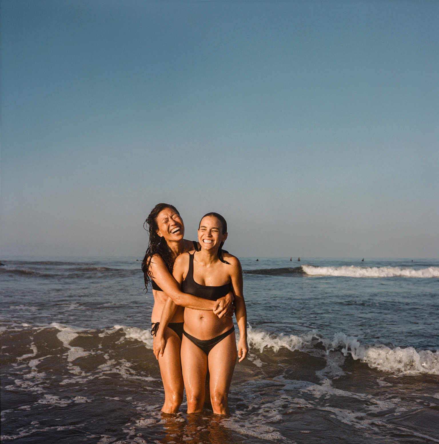 Two women in black bikinis embracing and laughing whilst standing in shallow ocean water with waves and blue-green sky behind them.