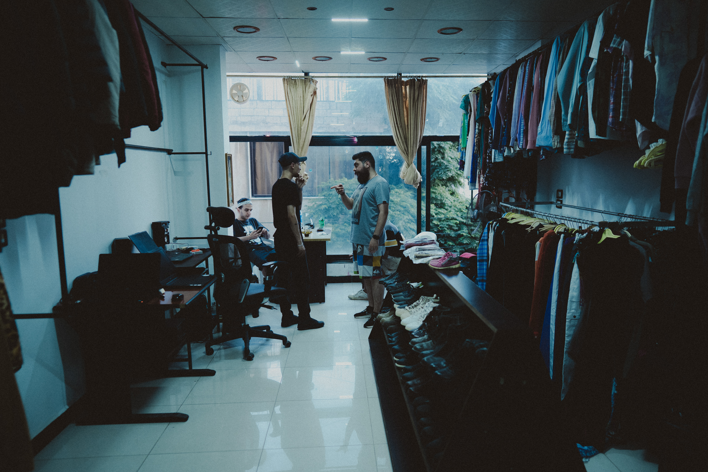 Interior of clothing shop with two people standing amongst racks of garments, white floors, fluorescent lighting, and blue-tinted atmosphere.