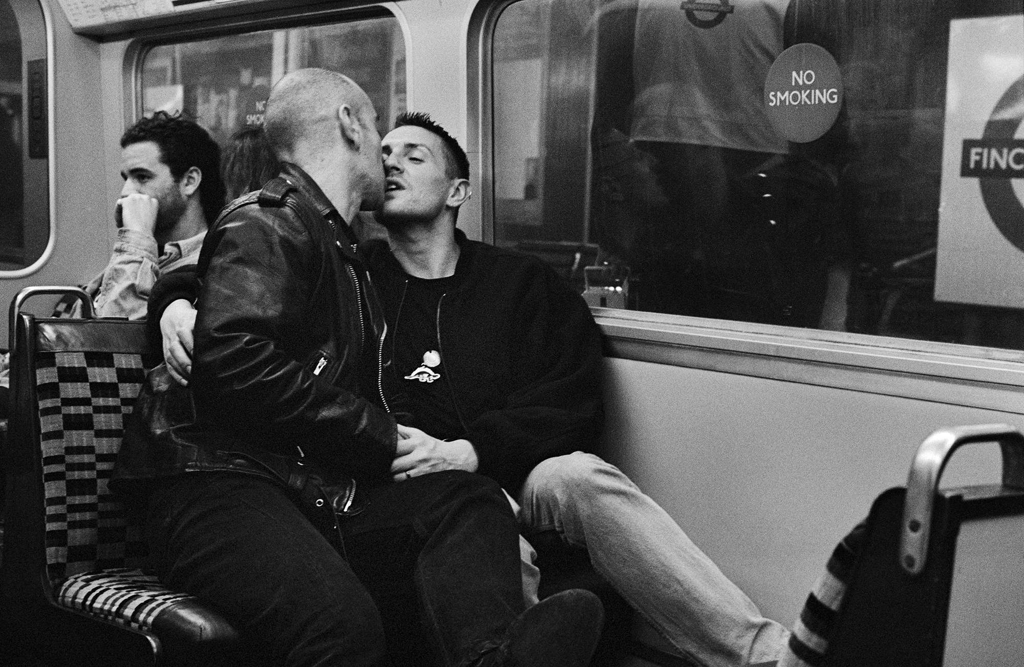 Black and white image of two men kissing on train seats, with another passenger visible in background near windows with "No Smoking" sign.