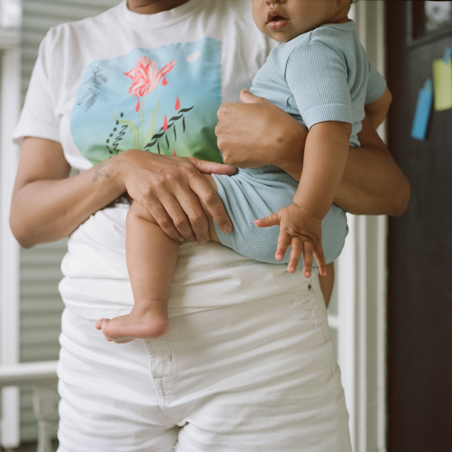 Person in white t-shirt with floral design holding toddler in light blue outfit, both wearing white trousers.