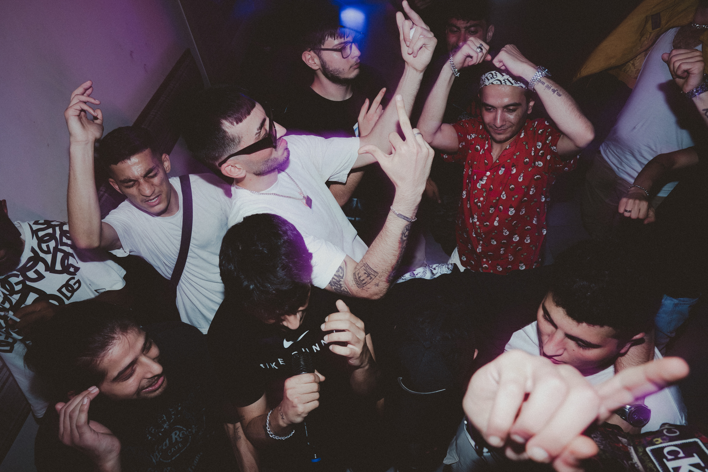 Group of young people dancing at nightclub with raised arms, purple and blue lighting, crowded dance floor.