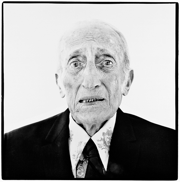 Black and white portrait of elderly man in dark suit and tie, with wrinkled face and thinning hair, against plain background.