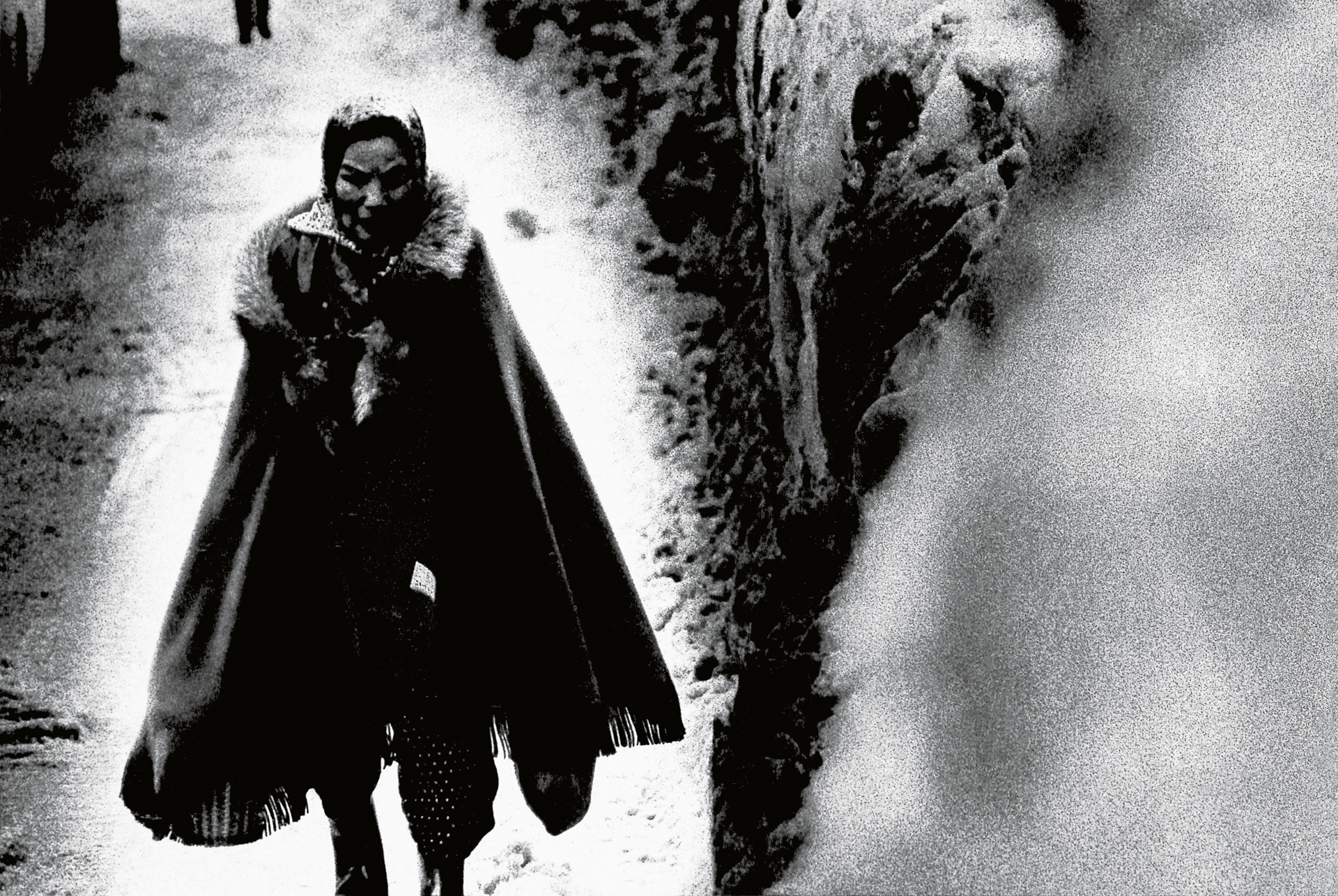 Black and white film still showing person in dark cloak walking through snowy courtyard with stone walls and archways.