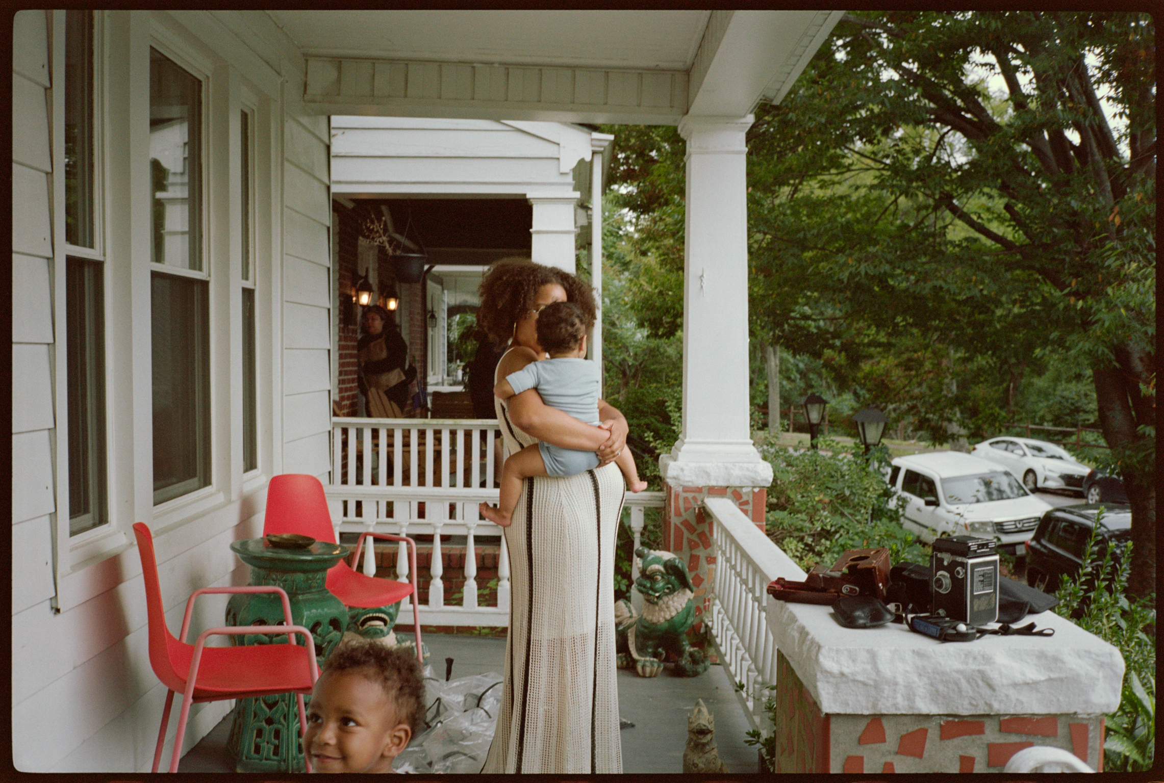 Woman holding child on white wooden porch with columns, red plastic chairs, white railings, green trees and parked cars visible.