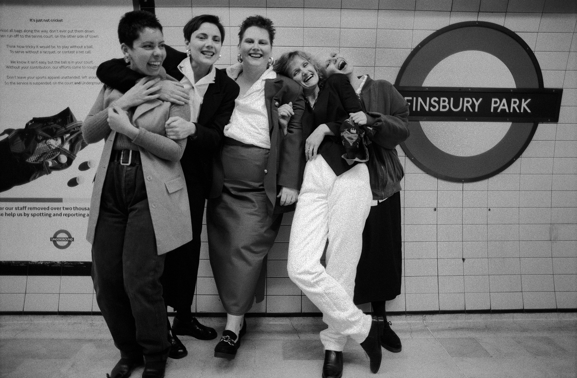 Five people embracing and laughing together on London Underground platform beside Finsbury Park station sign. Black and white image.
