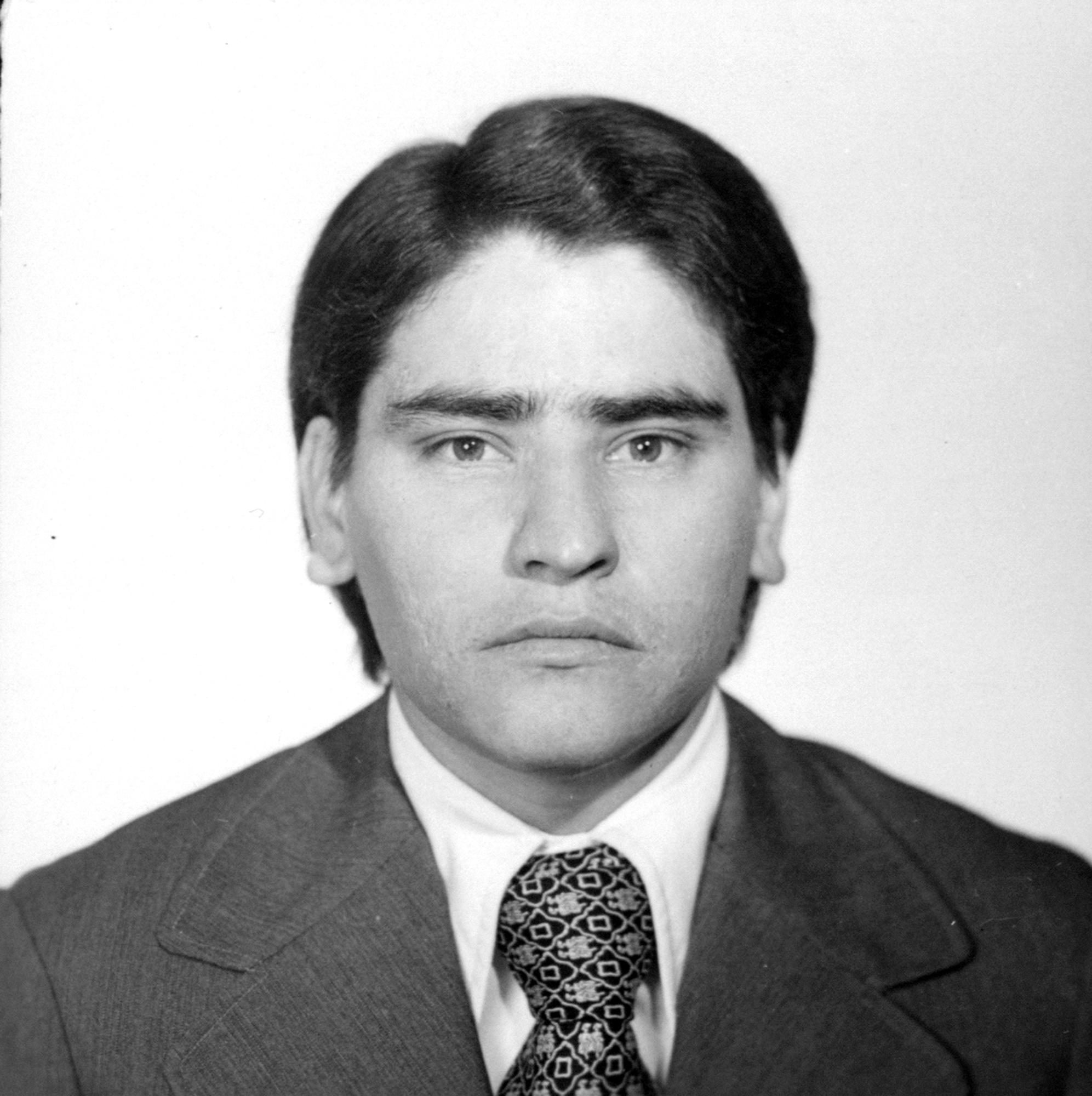 Black and white portrait of man with dark hair parted to side, wearing dark suit jacket, white shirt, and patterned tie.