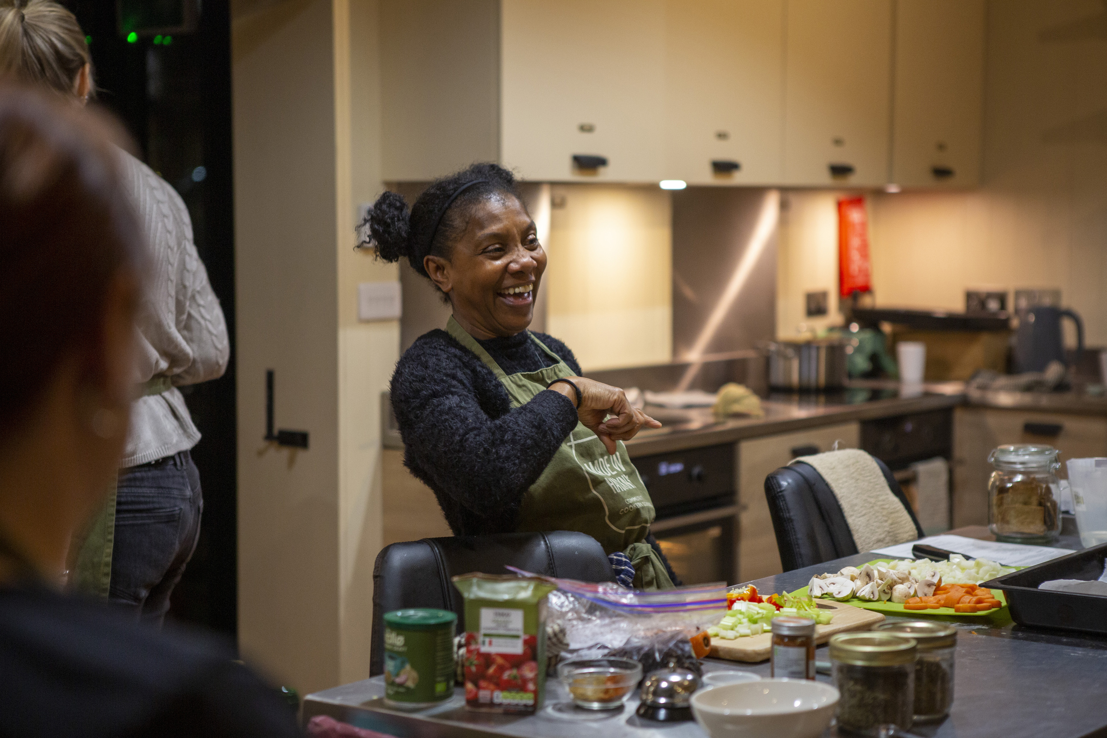 Woman cooking in modern kitchen with cream cupboards, smiling whilst stirring pot. Various ingredients and jars on counter.