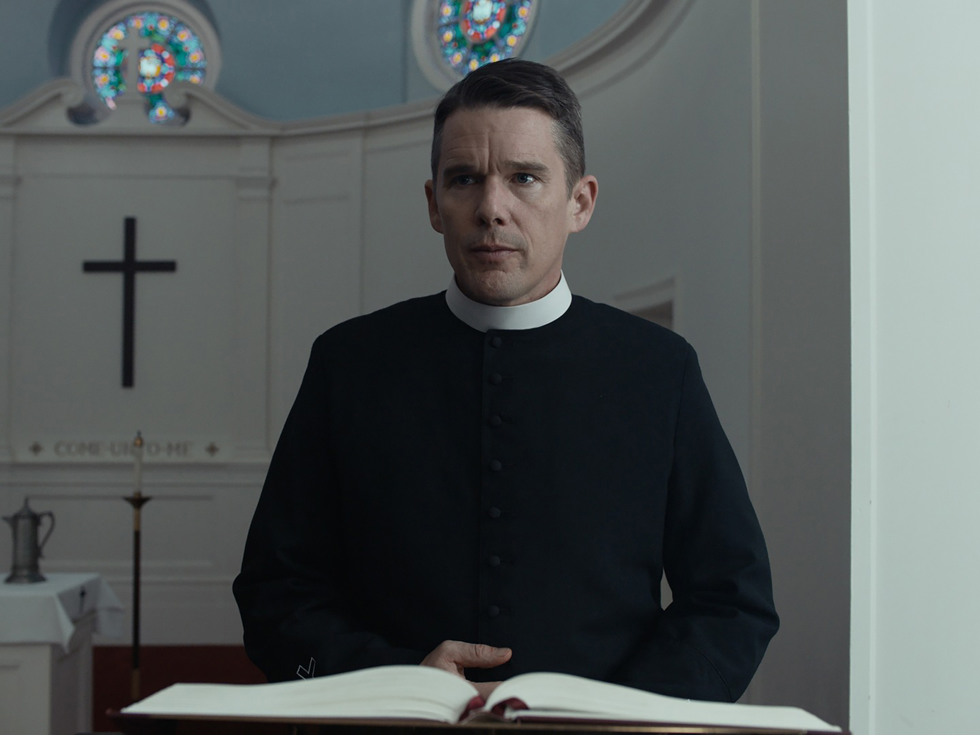 A man in a black clerical robe stands at a pulpit holding an open book.