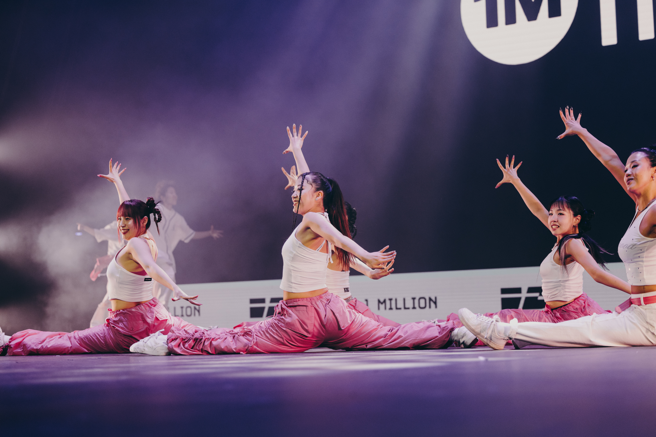 Four dancers in white tops and pink bottoms performing on stage with arms raised, purple lighting and "1M" signage visible in background.