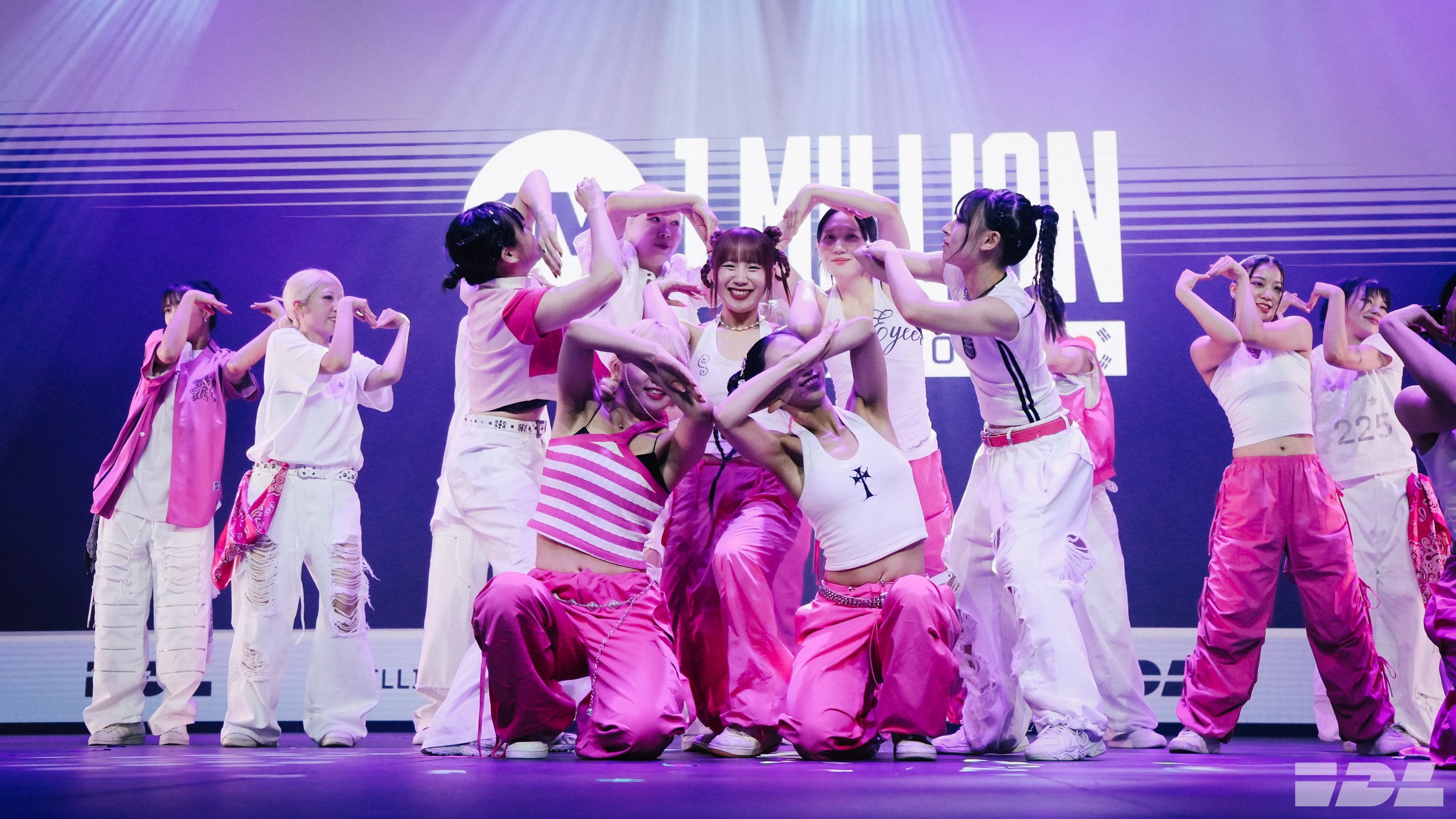 Group of performers in pink and white clothing embracing centre stage with purple and blue lighting backdrop showing "MILLION" text.