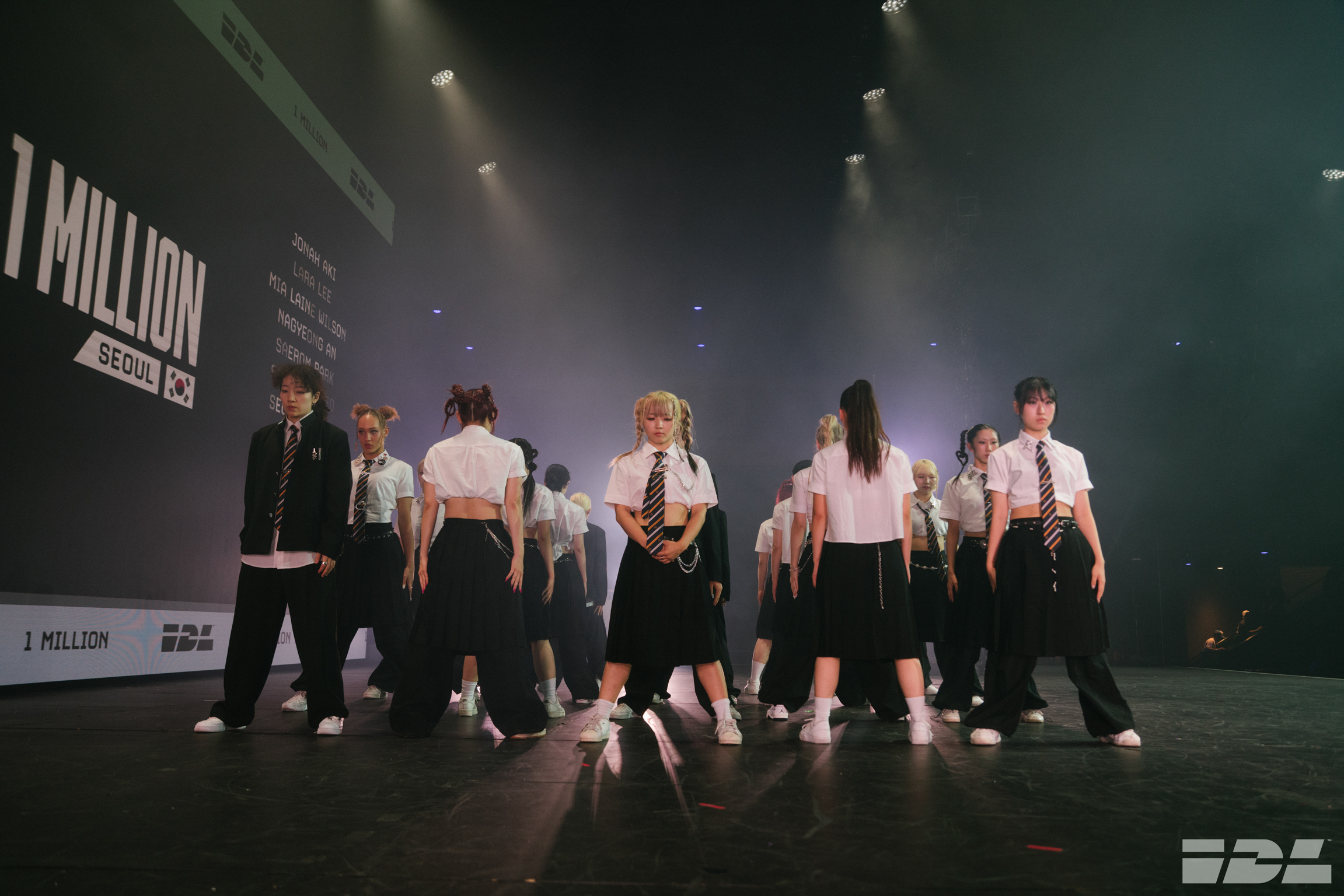 Group of performers in white shirts and dark trousers on stage with dramatic lighting and "J-MILLION" backdrop visible.