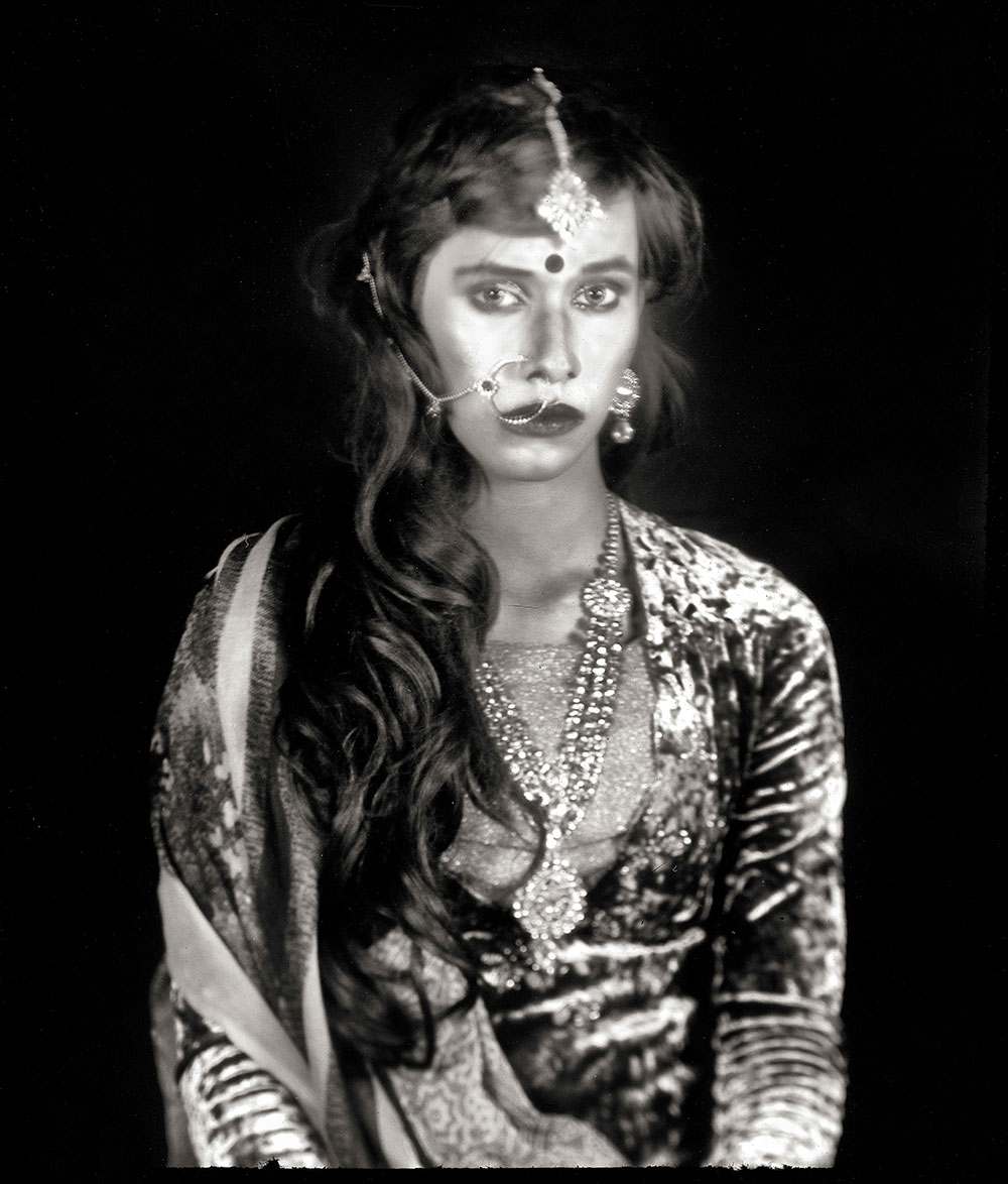 Woman in ornate Indian outfit with embellished blouse, jewellery including forehead piece and necklaces, long wavy hair, black background.