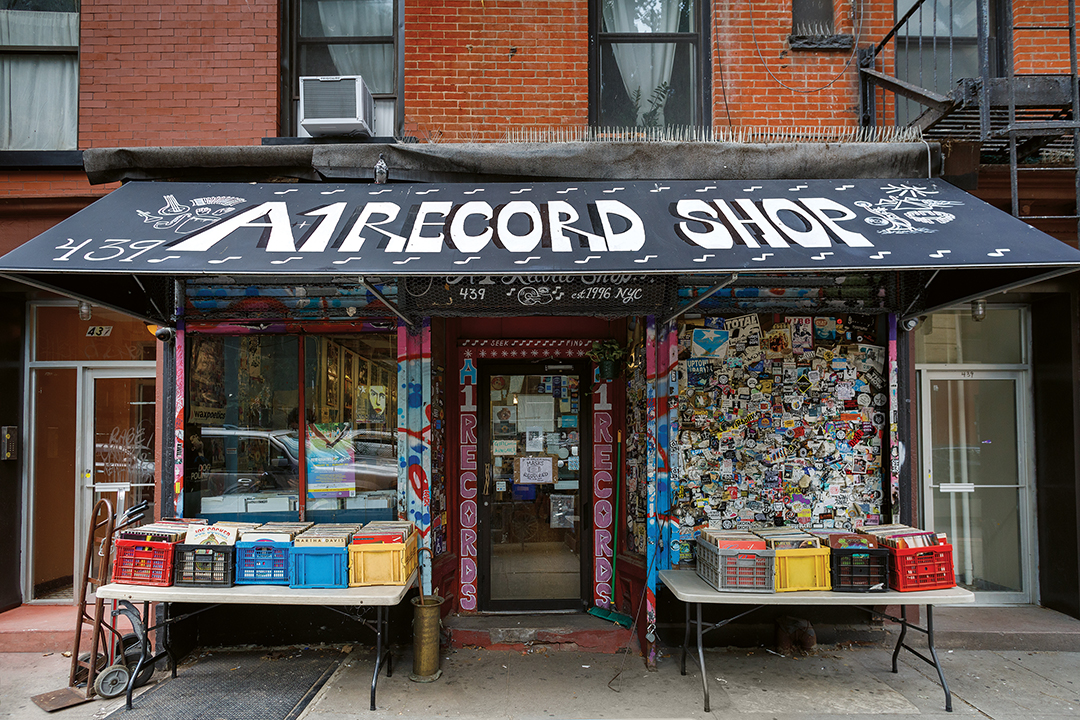 Record shop storefront with black awning, white text signage, coloured vinyl crates on tables, graffitied walls, red brick building above.