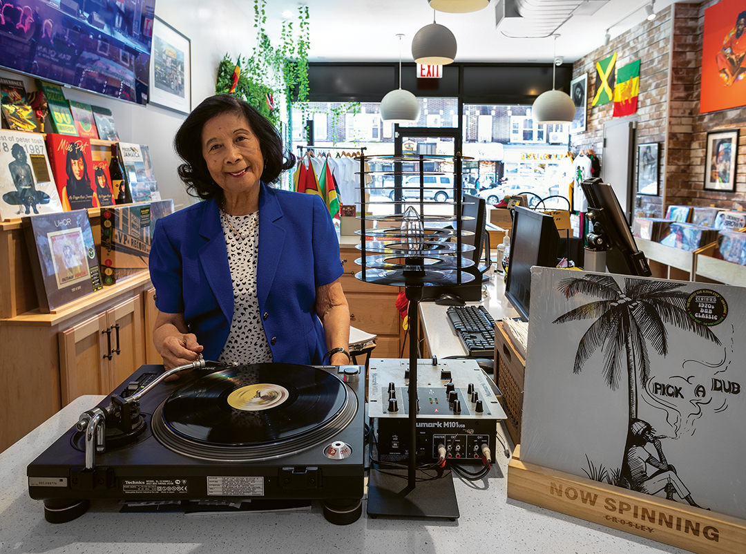 Woman in blue blazer standing behind turntable in record shop with vinyl records, artwork on walls, and shelving displays.