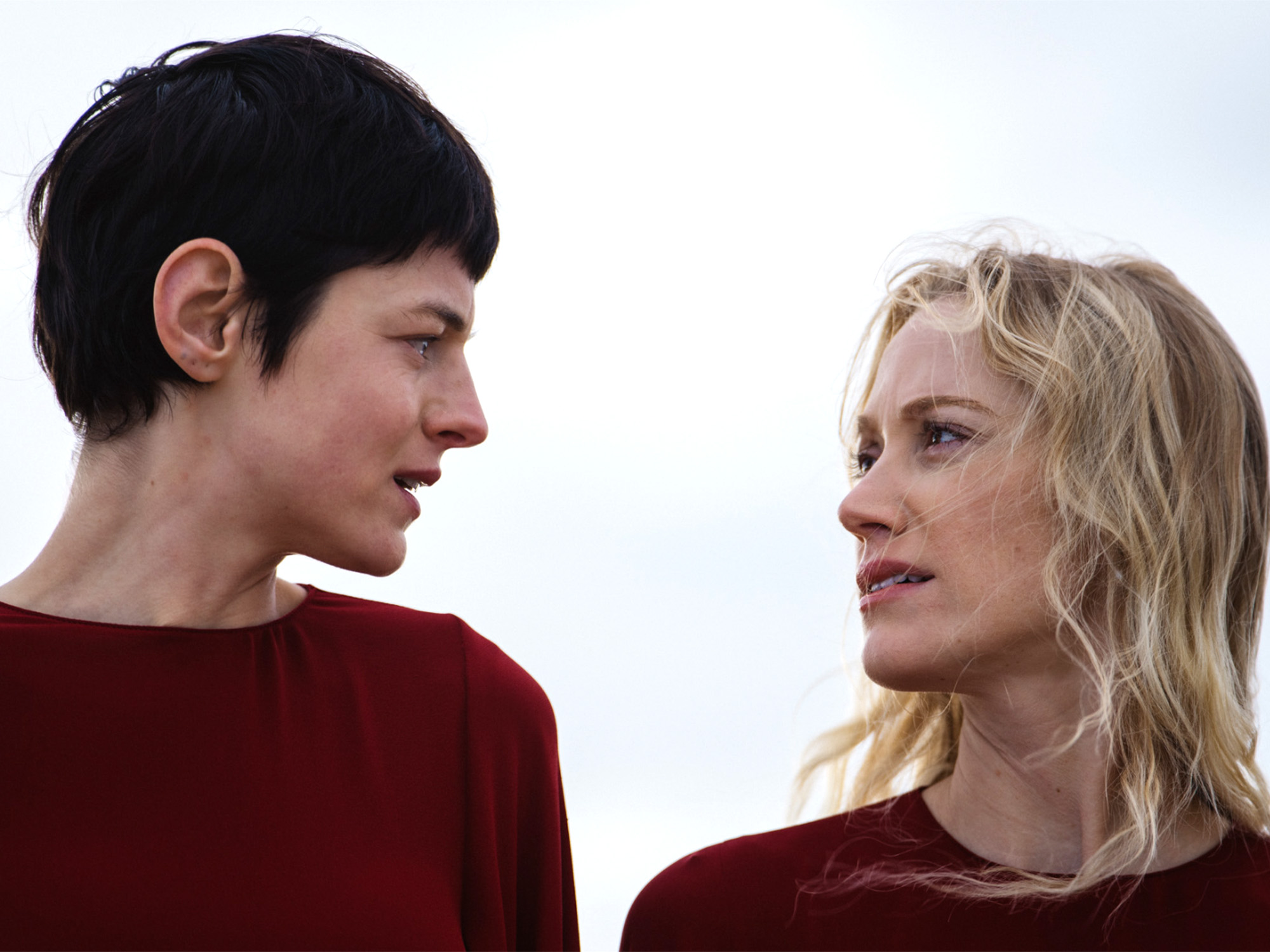 Two women in red tops facing each other against cloudy sky - one with short dark hair, one with blonde wavy hair.