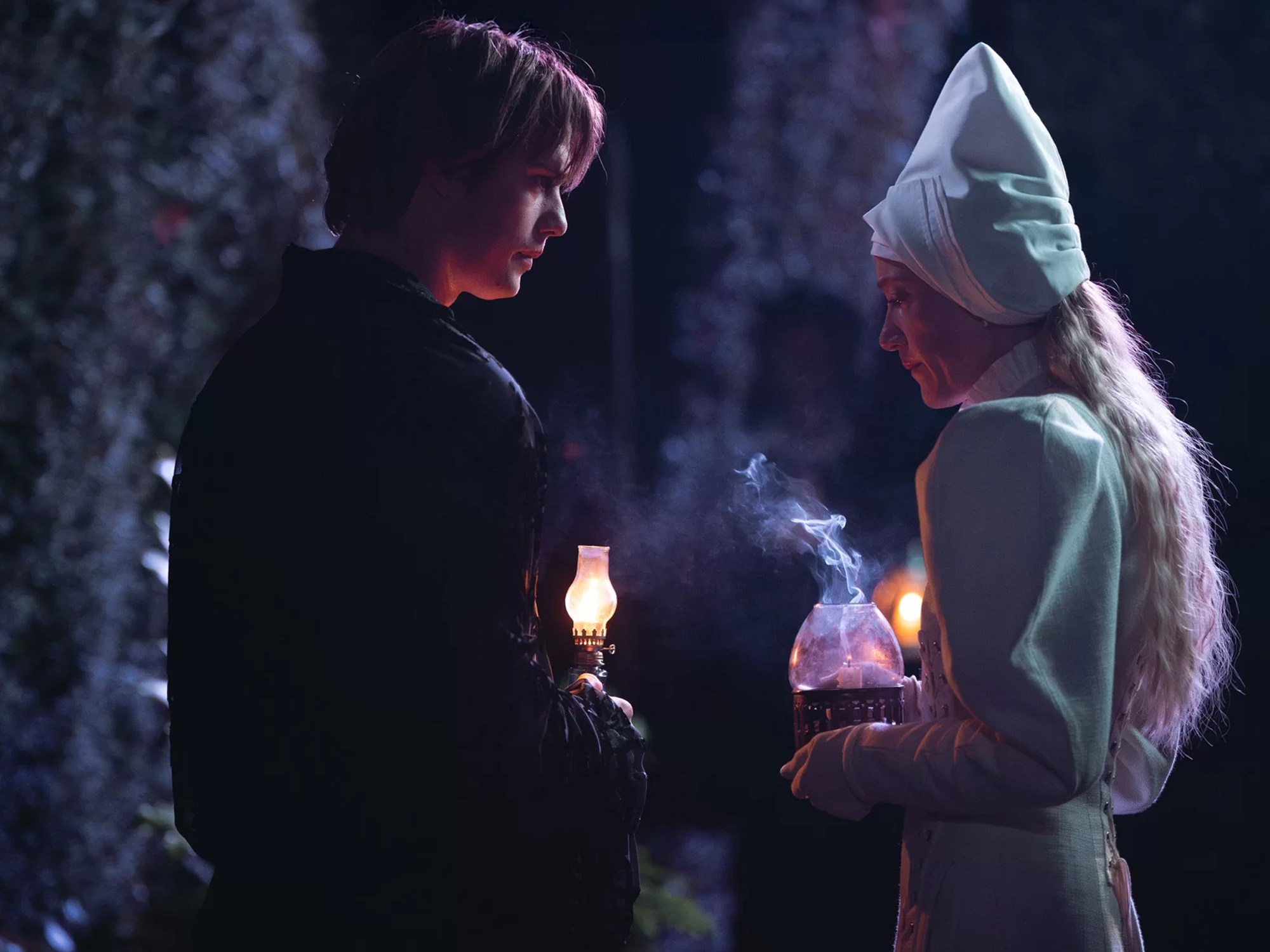 Two people in dark setting holding lit candles, one wearing white hooded garment, warm orange candlelight against cool blue background.