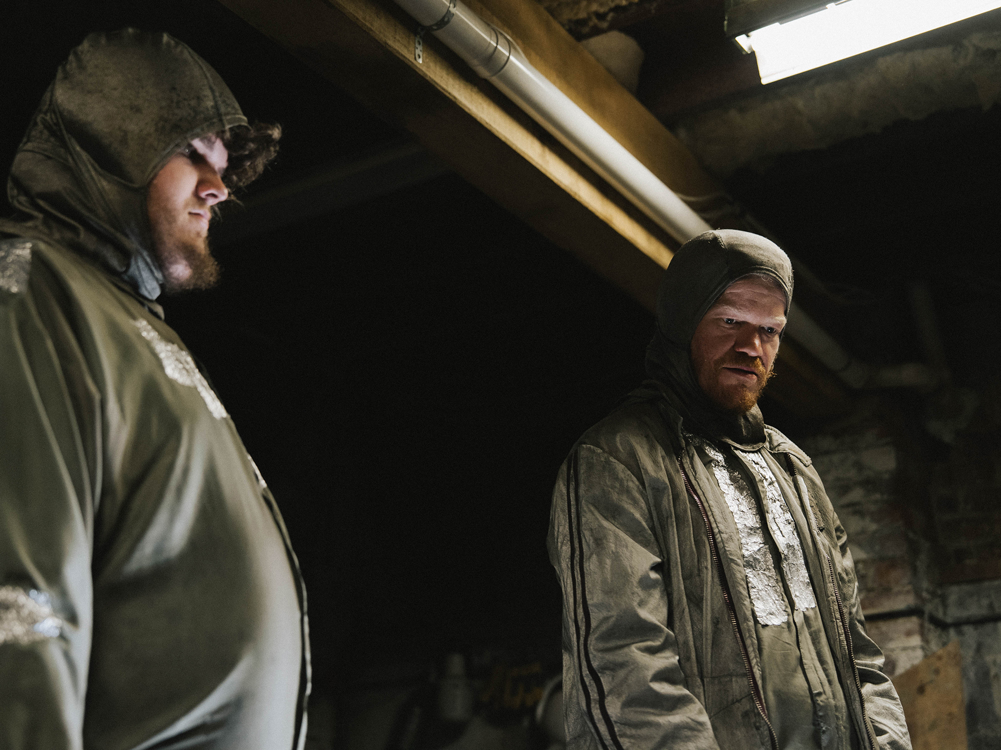 Two men in hazmat suits and gas masks standing in dark industrial basement with exposed pipes and fluorescent lighting.