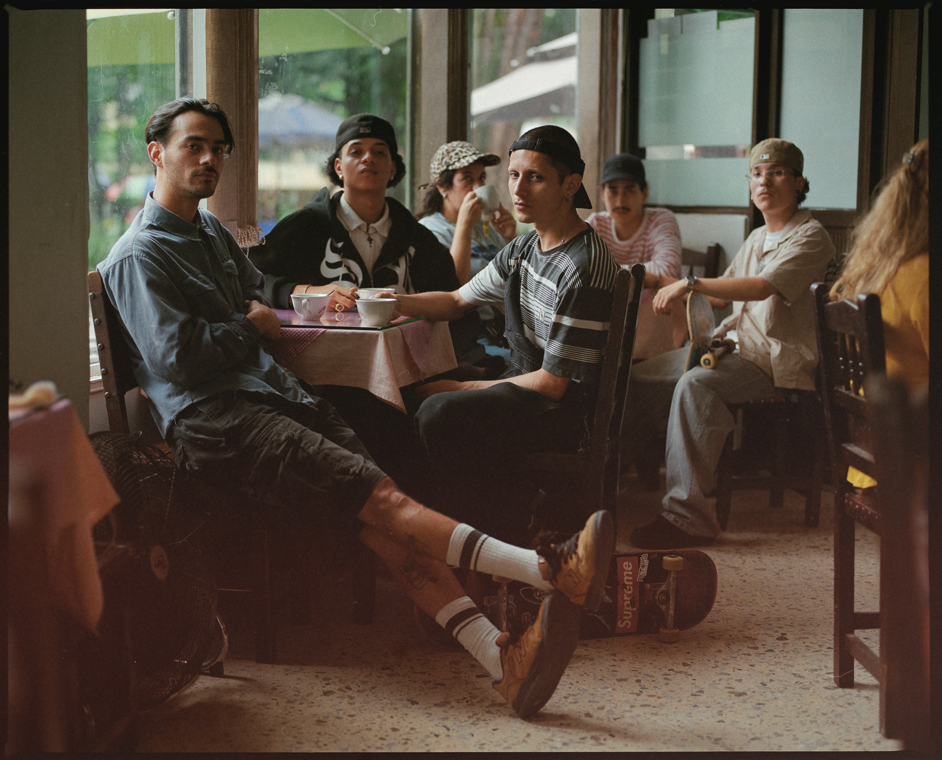 Group of people sitting at wooden tables in restaurant or café, large windows showing outdoor view in background.