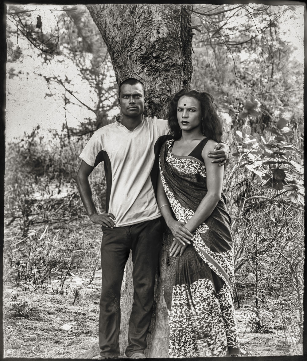 Black and white portrait of man and woman standing by tree trunk, he in t-shirt and trousers, she in patterned dress.
