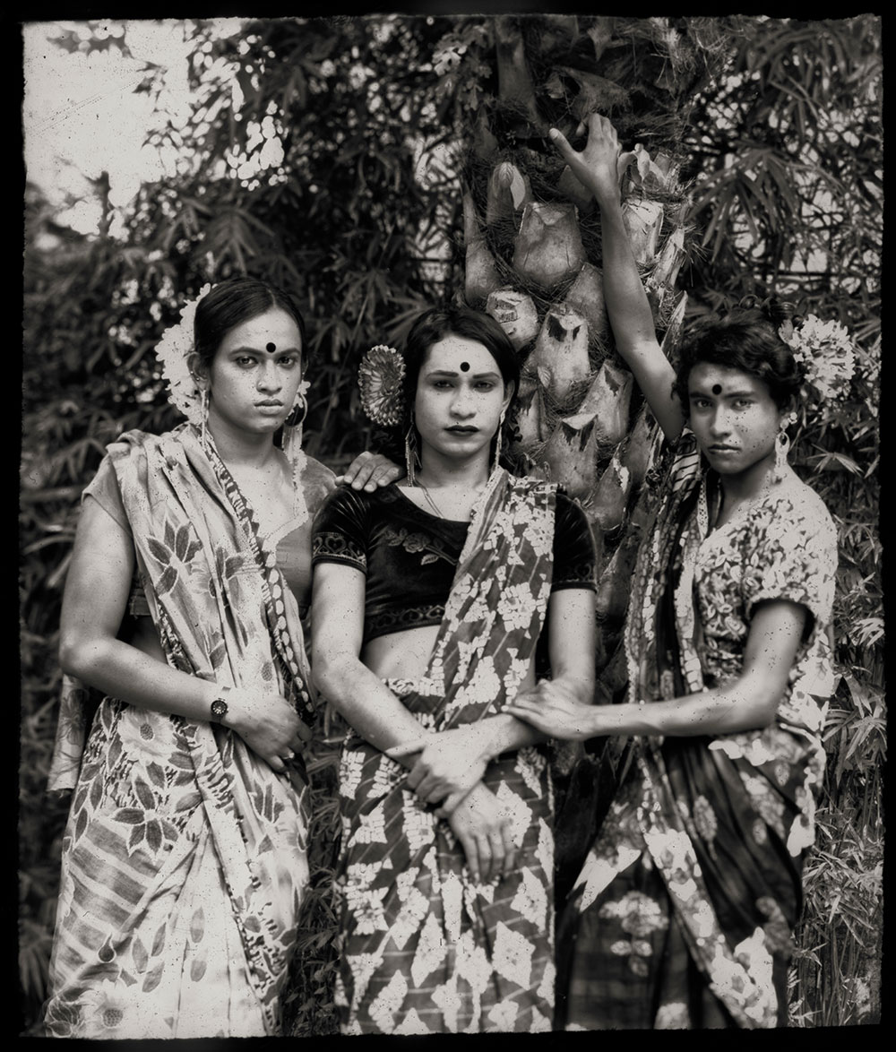 Three women in traditional Indian saris and jewellery stand together outdoors. Black and white photograph with foliage background.