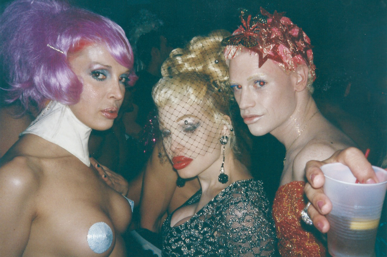 Three women in elaborate costumes and makeup, posing together against a dark background.