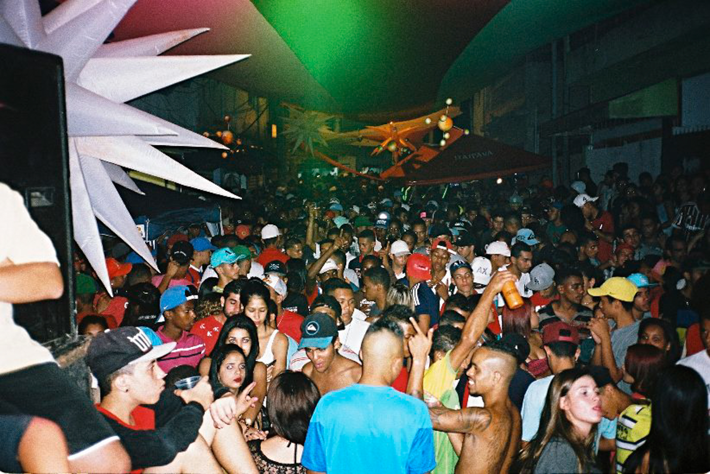 Crowded nightclub interior with densely packed people, green lighting overhead, white decorative elements on left wall.