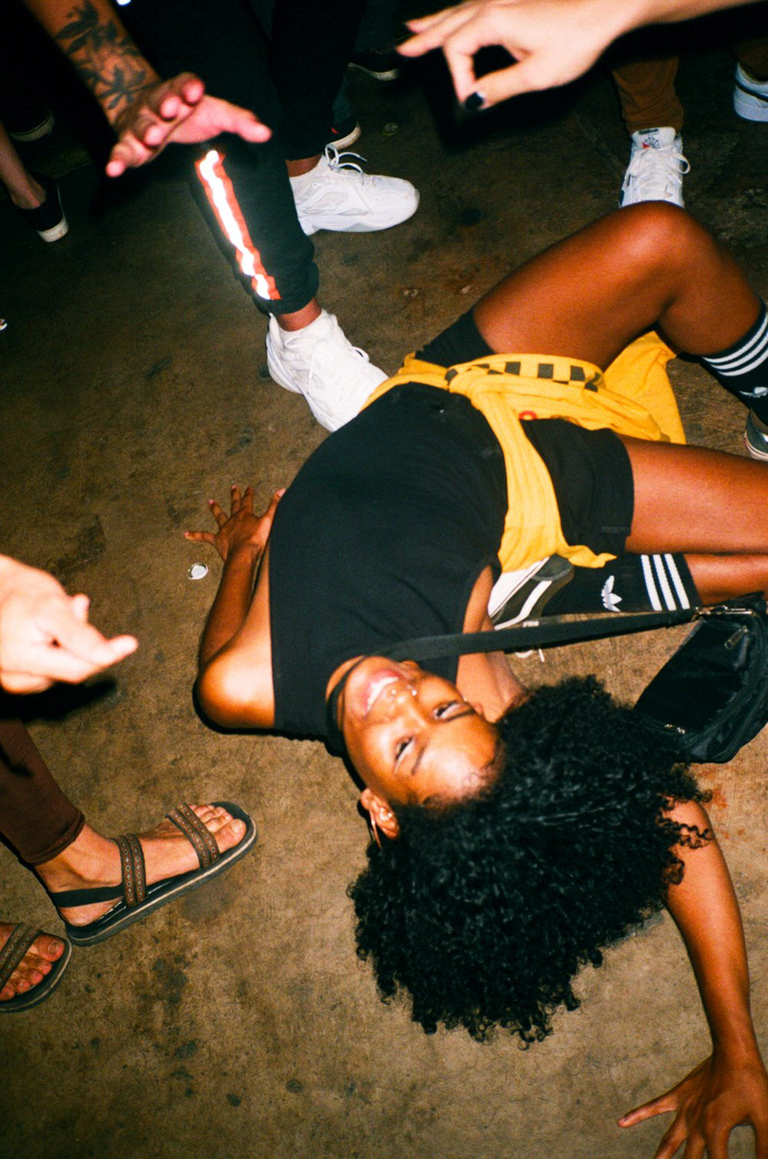 Person with curly dark hair lying on concrete floor wearing yellow sports bra and black shorts, surrounded by feet in trainers and sandals.