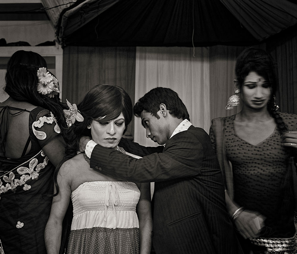 Man in dark suit adjusting woman's strapless dress whilst other women in traditional clothing look on indoors.