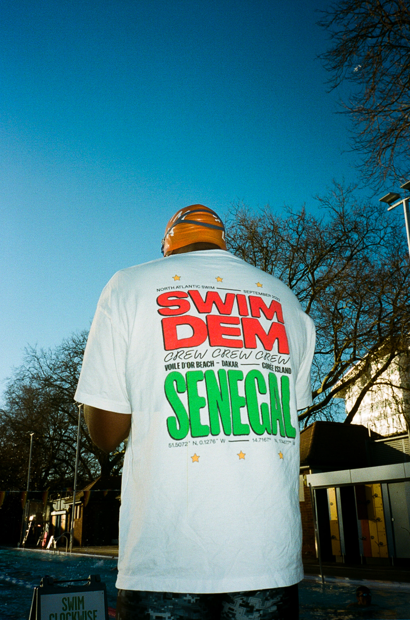 Person wearing orange beanie and white t-shirt with red "SWIM DEM" and green "SENEGAL" text, viewed from behind against blue sky.