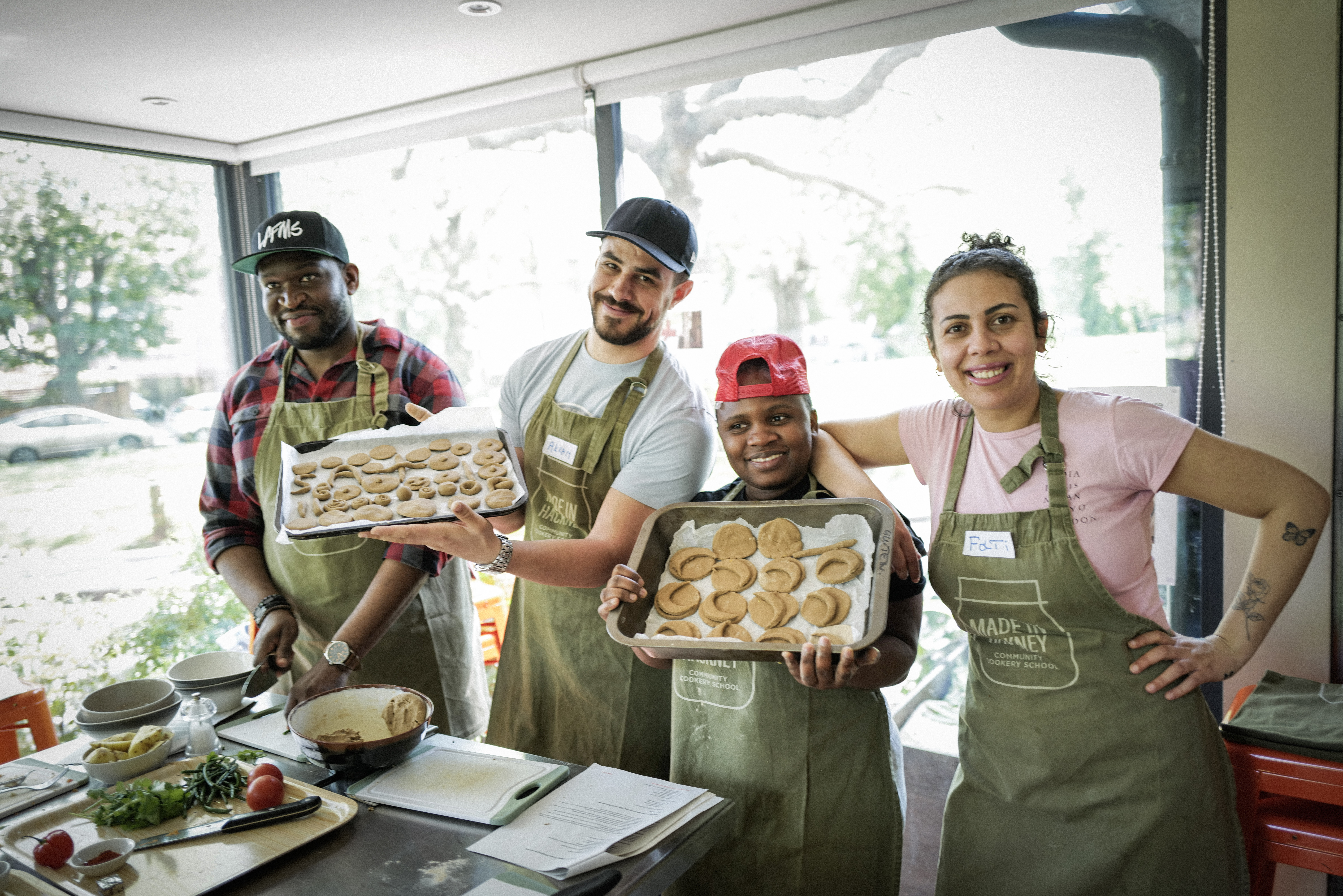 Four kitchen staff in green aprons and caps holding baking trays of pizza and bread rolls, smiling in commercial kitchen with large windows.