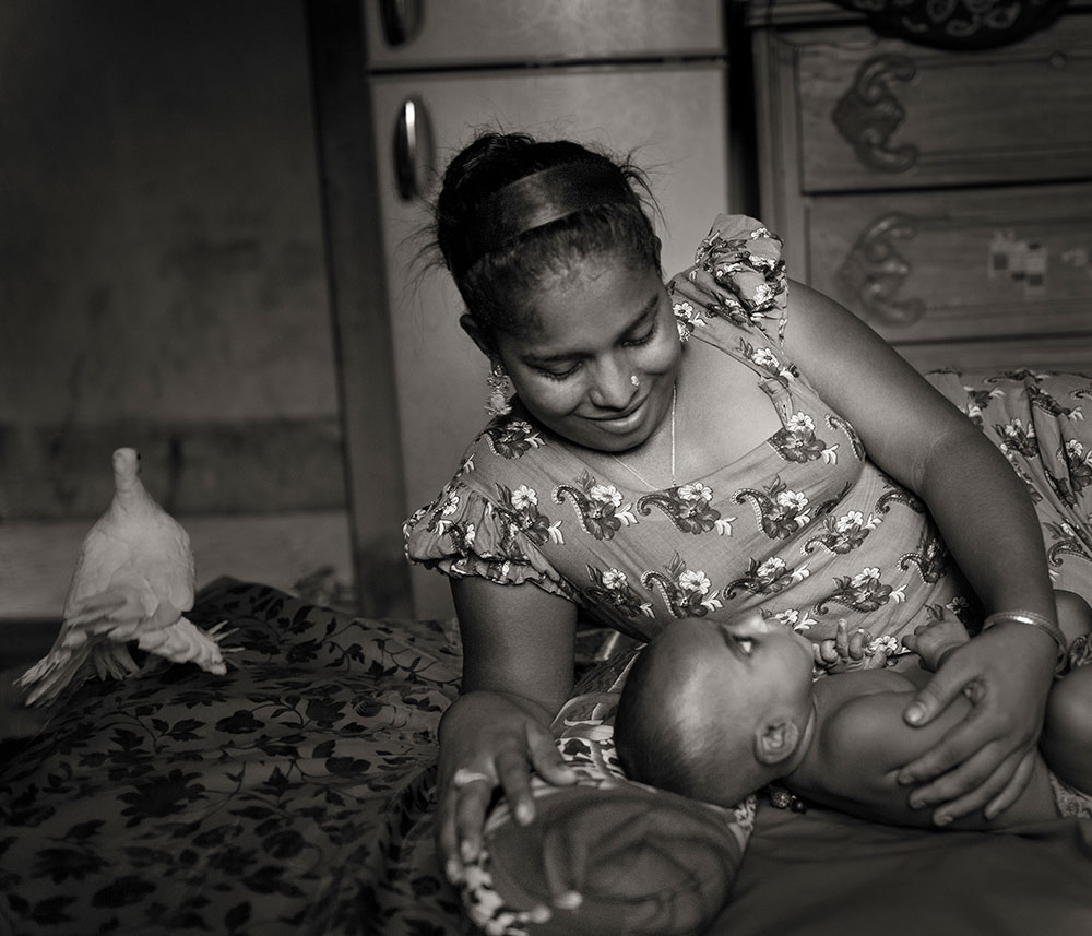 Woman in floral dress breastfeeding infant whilst sitting on patterned fabric indoors, with framed pictures on wall behind.