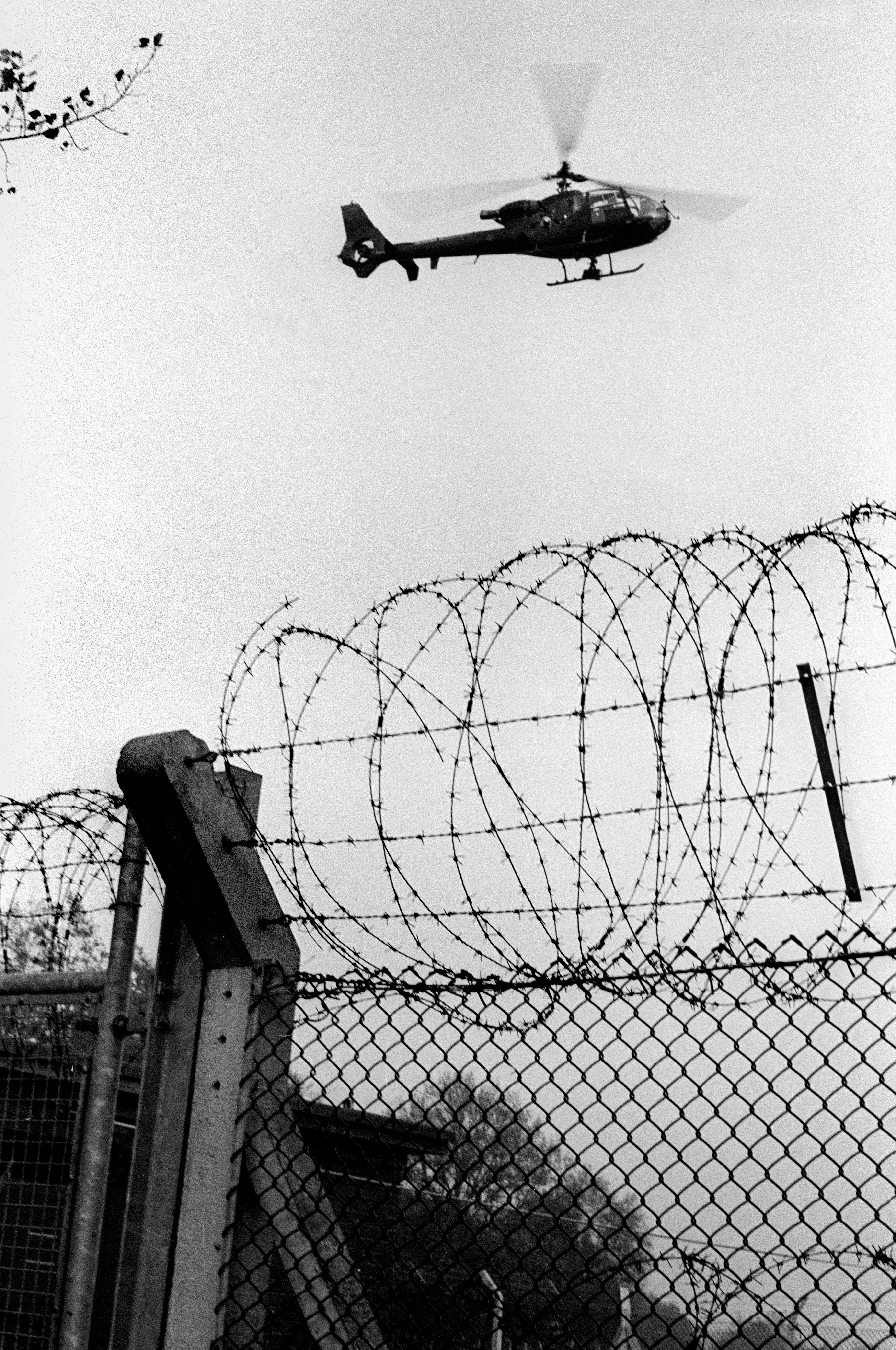 Black and white image: helicopter flying above razor wire-topped chain-link fence with concrete post in foreground.