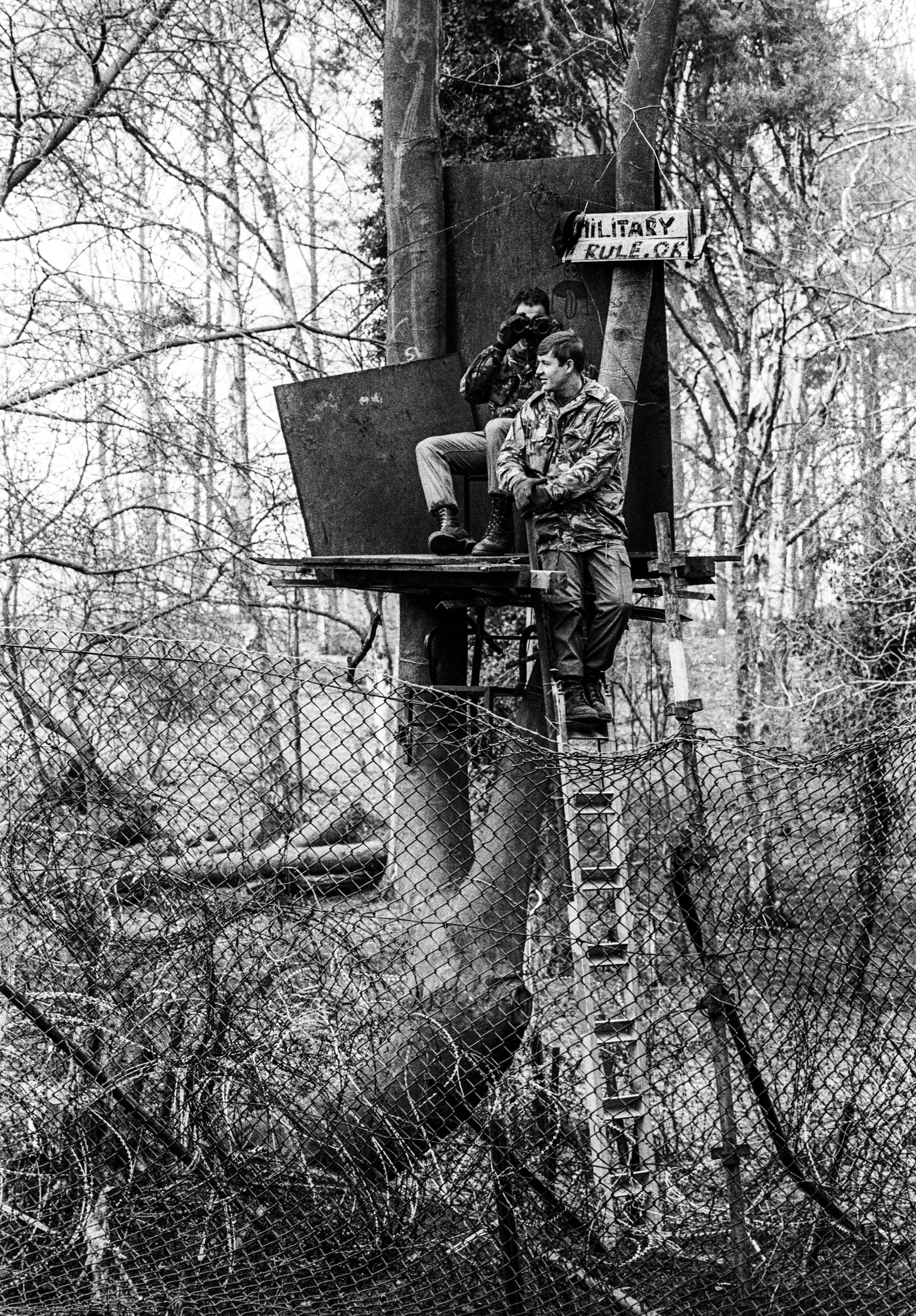 Black and white image of military personnel in elevated observation post with ladder, surrounded by bare trees and chain-link fencing.