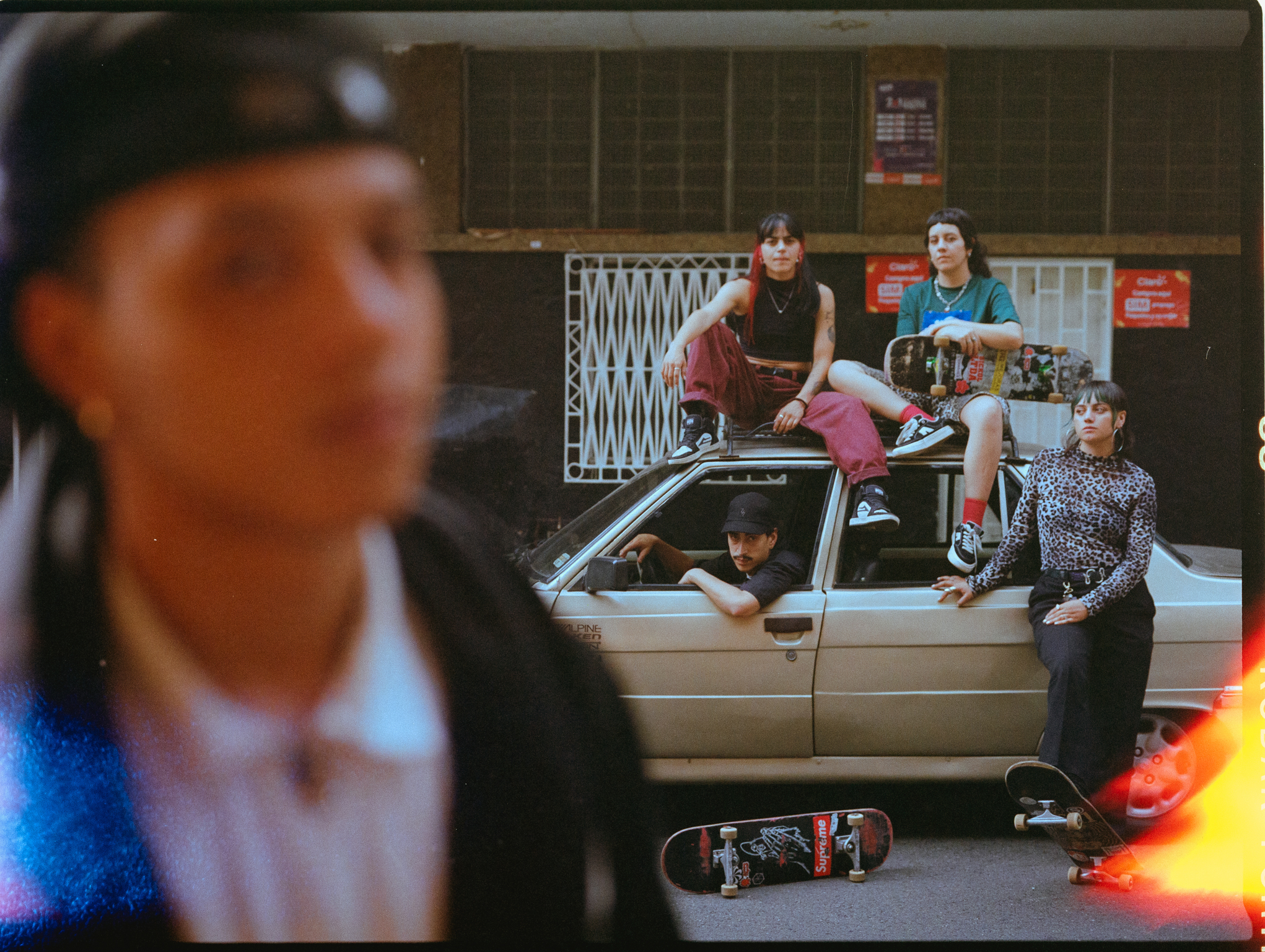 Group of young people gathered around white car on street, with blurred figure in foreground and orange light visible at bottom right.
