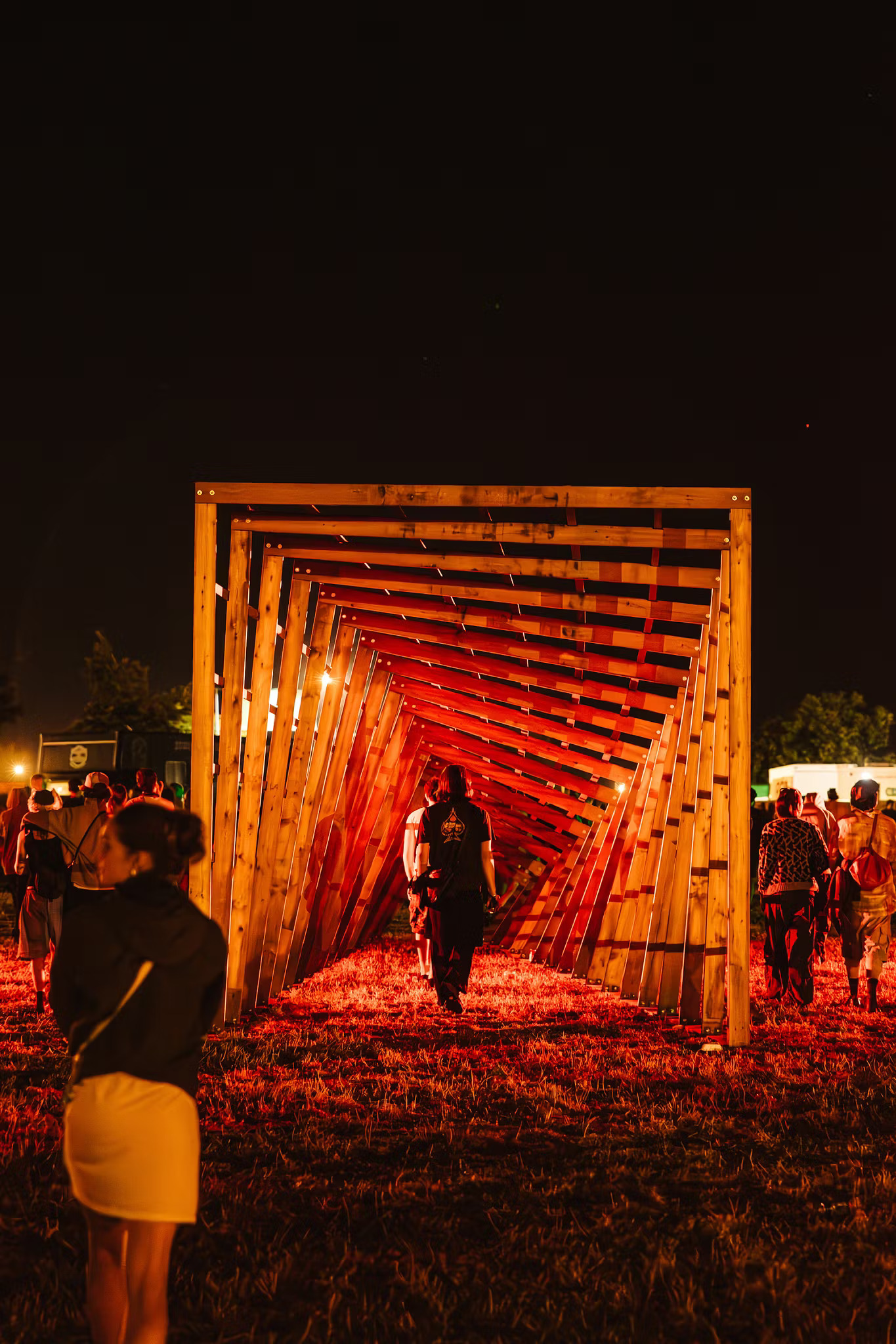 Illuminated wooden tunnel structure with orange and red lighting at night, people walking through and around it on grass.