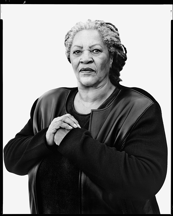 Black and white portrait of Toni Morrison seated, wearing dark clothing with arms crossed, looking directly at camera.