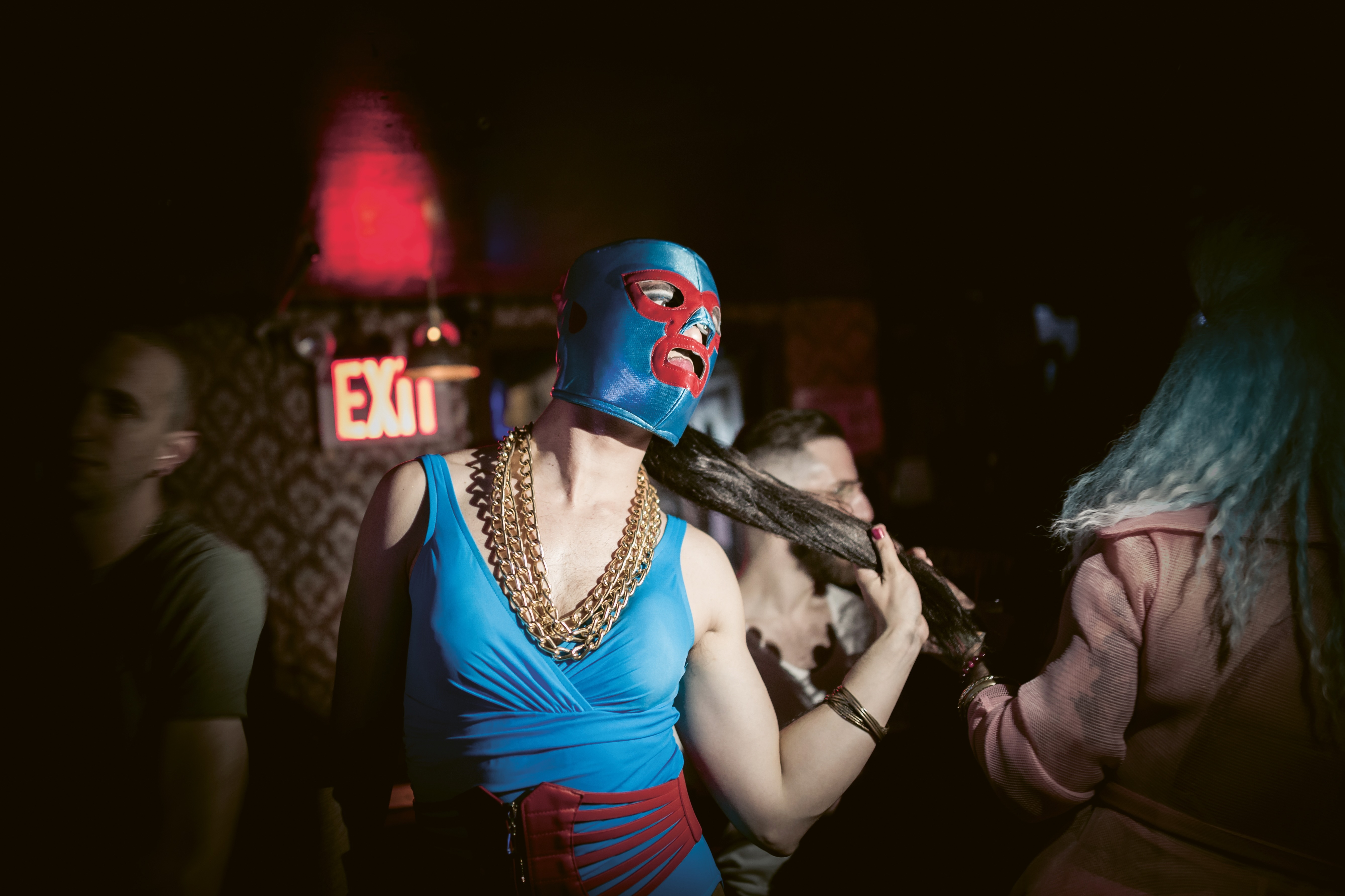 A person wearing a blue and red face mask, gold necklaces, and a blue top performing on stage at night with an exit sign visible in the background.