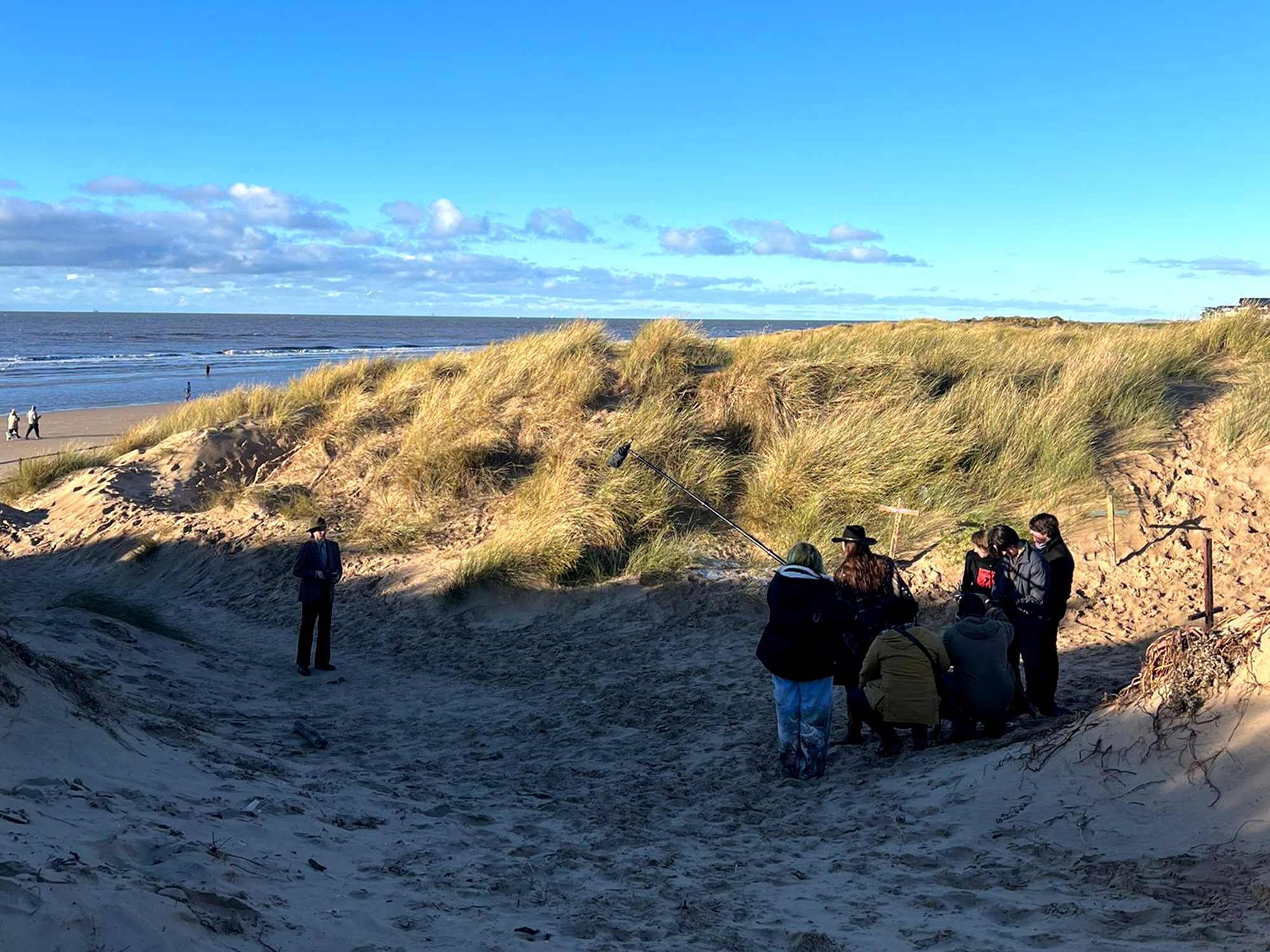 Sandy beach with golden grass-covered dunes, people gathered near wooden steps, blue sea and cloudy sky in background.