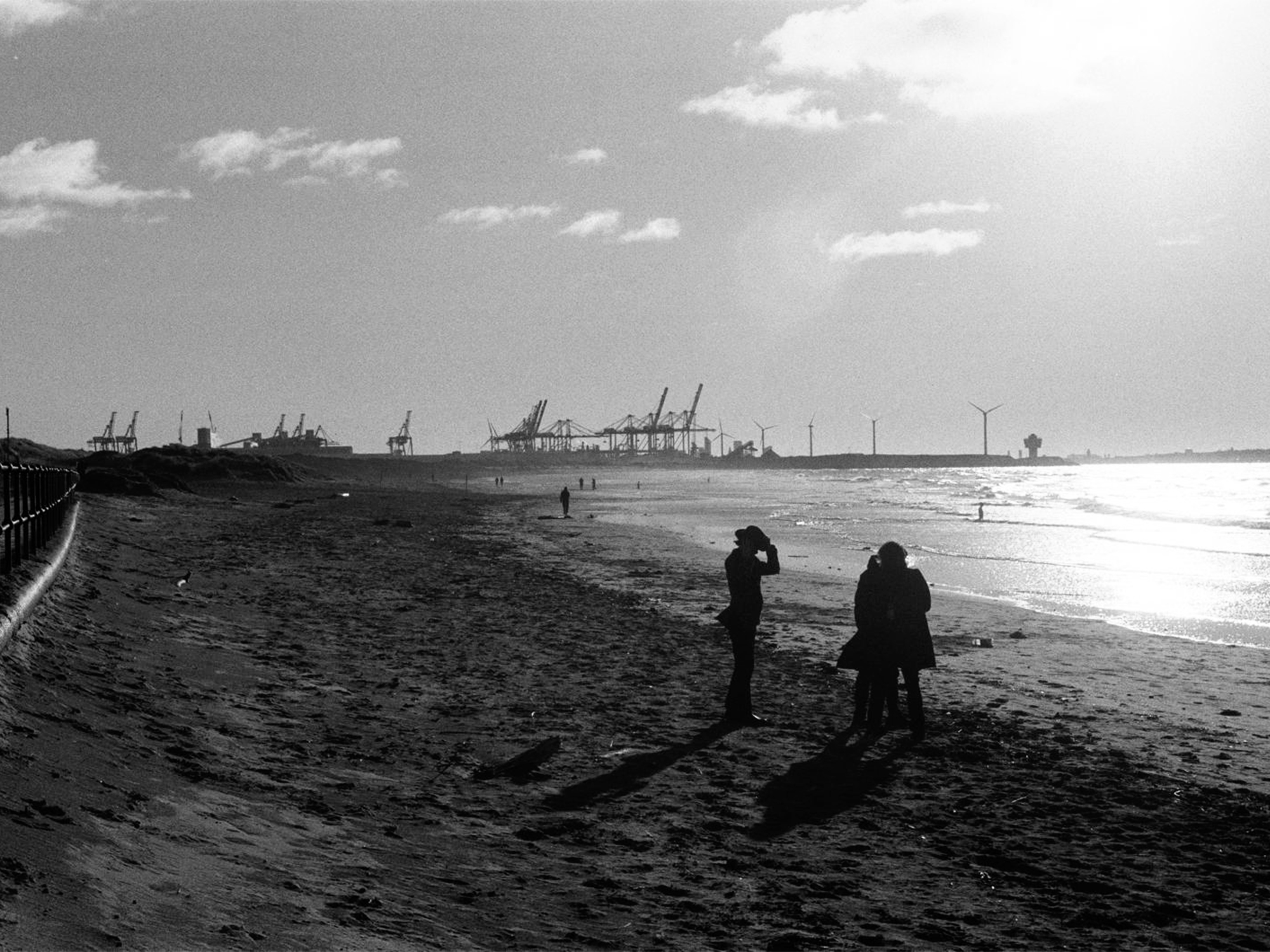 Black and white image of people on sandy beach with industrial port cranes visible on horizon under cloudy sky.