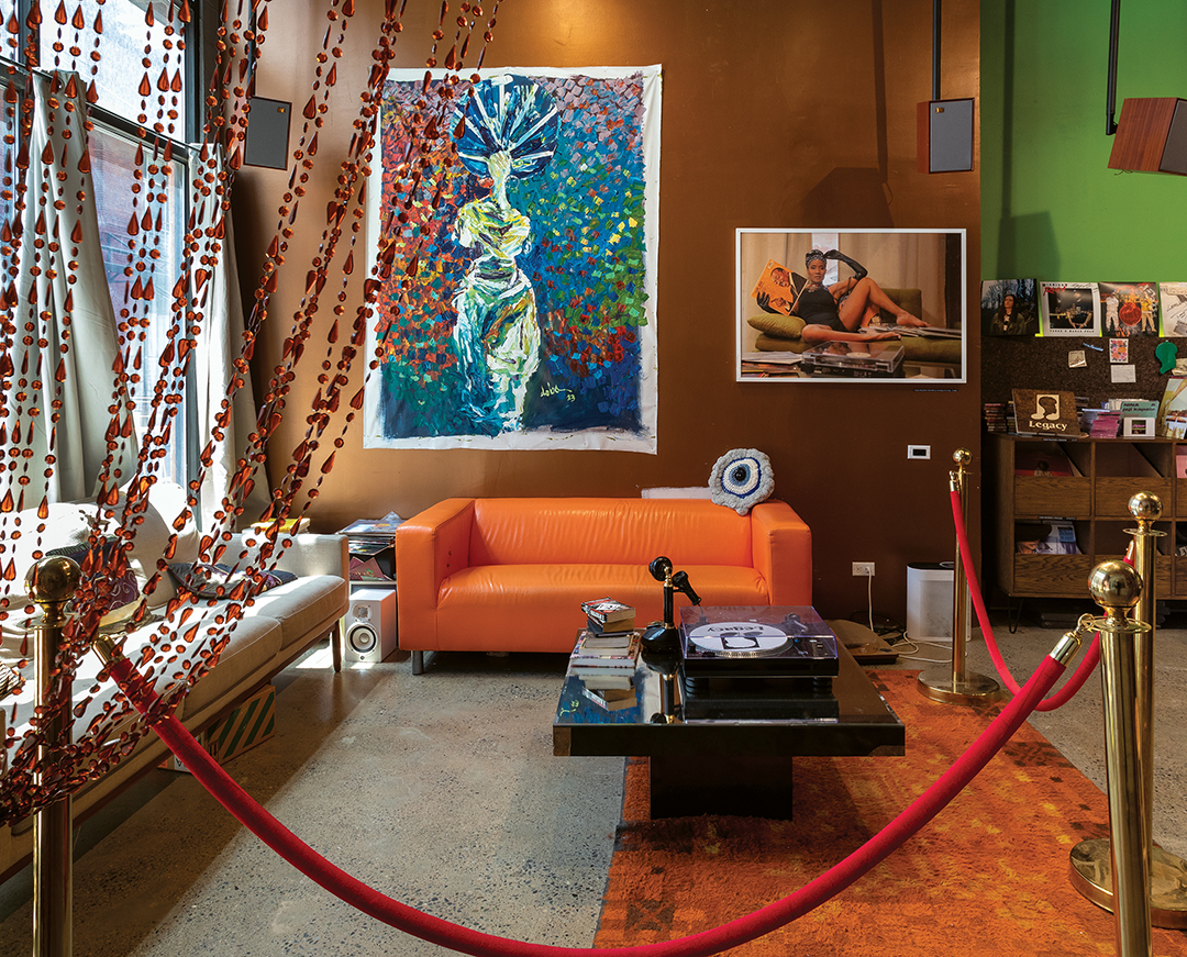 Gallery space with red velvet rope barrier, orange sofa, patterned curtains, and blue-toned artwork on green walls.